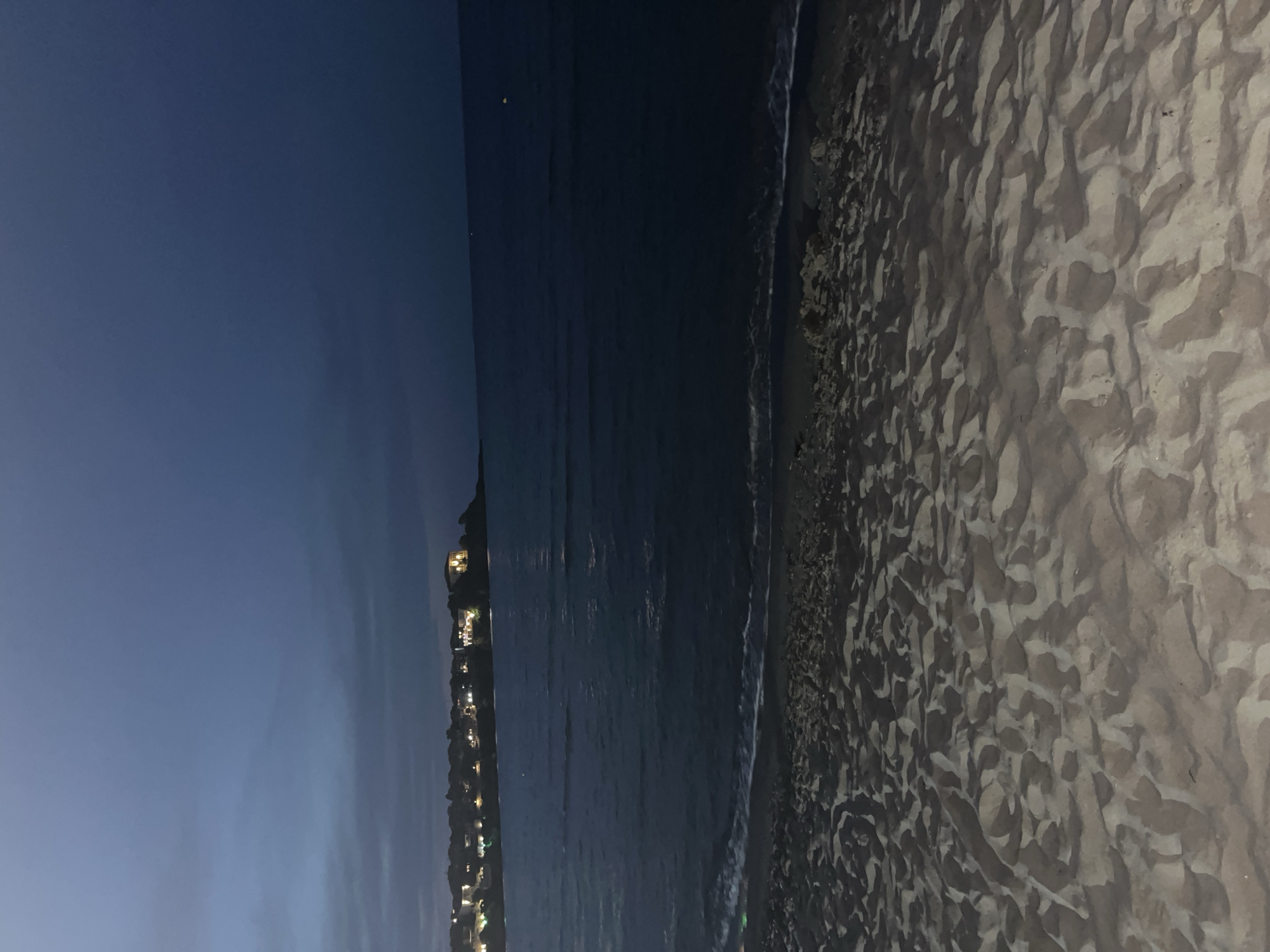 The image depicts a twilight scene on a sandy beach in Sozopol, Bulgaria. The sky is a deep, dark blue, transitioning to a lighter shade near the horizon. In the distance, a coastline is visible, dotted with illuminated buildings that suggest a town or city. The sea is dark and calm, with gentle waves lapping at the shore. The foreground is dominated by the textured surface of the sand, which is illuminated by an unseen light source, creating patterns of light and shadow. The overall impression is one of a peaceful evening by the sea in Sozopol.