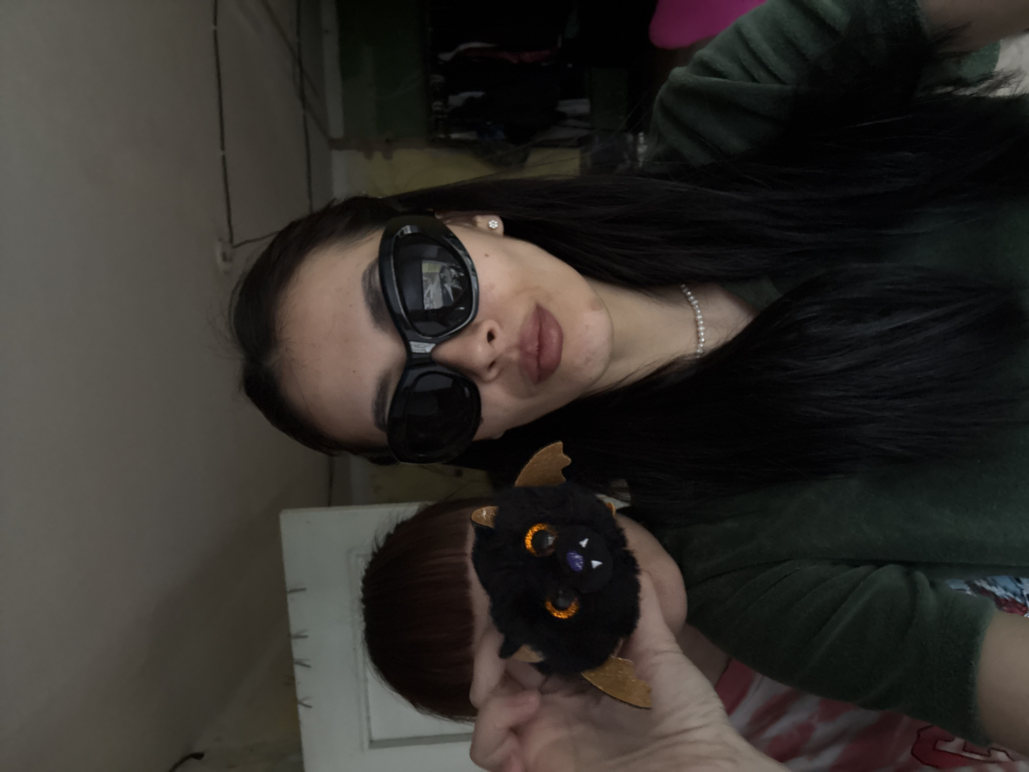 A selfie of a woman in an indoor setting, possibly a residence. The woman is wearing large, black sunglasses and a green, velour top. A pearl necklace is visible around her neck, and she has an earring. She's holding a small, black, fuzzy bat toy with golden wings. Another person's head is partially visible behind the bat toy. The location is purported to be Votkinsk, Russia, but there's no visual confirmation of the city. The background is slightly cluttered and suggests a lived-in space, but specific details are obscured.