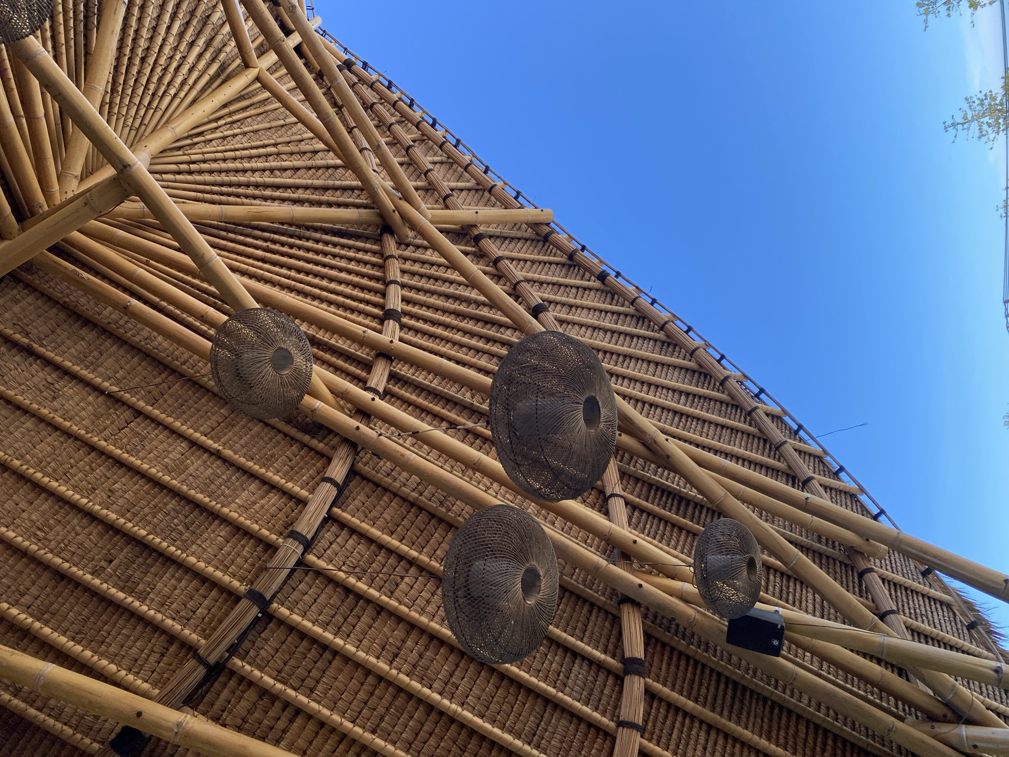 A low-angle shot showcases the intricate bamboo and thatch roof of a structure, with a clear blue sky visible in the upper right. Four dark-colored, spherical lamps or decorative objects hang from the roof's framework. The structure appears to be located in Kuta Selatan, Indonesia.