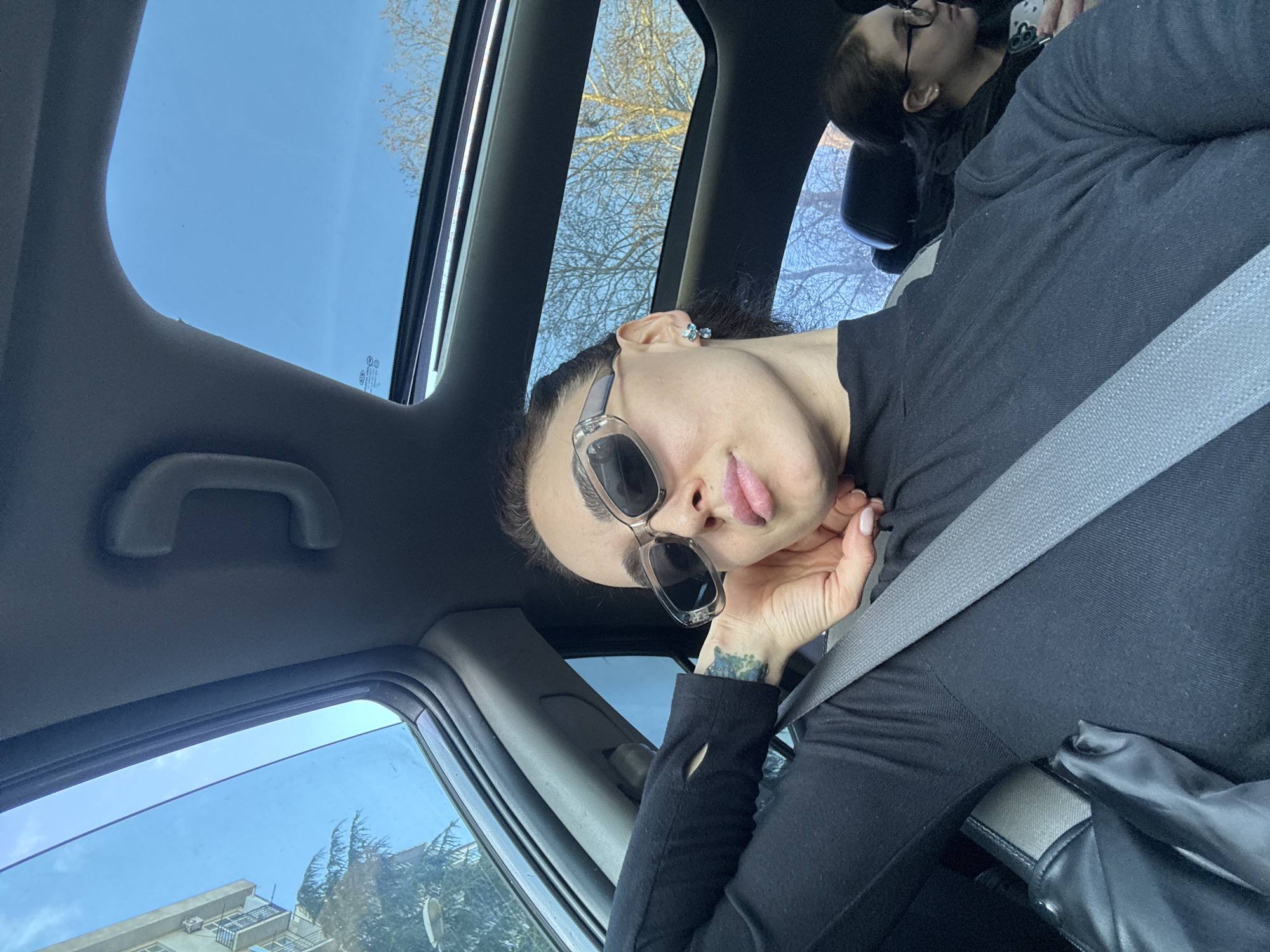The image depicts the interior of a vehicle located in Tbilisi, Georgia, during daytime under a clear blue sky. Two female individuals are present.
The primary subject, seated in the front passenger seat, is a female with dark hair, wearing clear-framed sunglasses with dark lenses and a black long-sleeved turtleneck shirt. Her right hand is positioned with fingers resting against her jawline/chin; her nails are manicured with light polish. A blue/green tattoo is partially visible on her left forearm. She is wearing small blue gemstone earrings. A gray seatbelt diagonally crosses her torso. A black bag is visible on her lap.
A second female individual is partially visible in the rear seat behind the primary subject. She wears eyeglasses with dark frames and has dark hair. Her right hand is holding a smartphone with a light-colored case.
The vehicle's interior features a gray headliner and a panoramic sunroof, through which a clear blue sky and deciduous trees with bare branches are visible. A grab handle is present above the front passenger door. Through the front left window, a multi-story residential building with balconies and multiple external air conditioning units is seen. A street light fixture is also visible in the background.