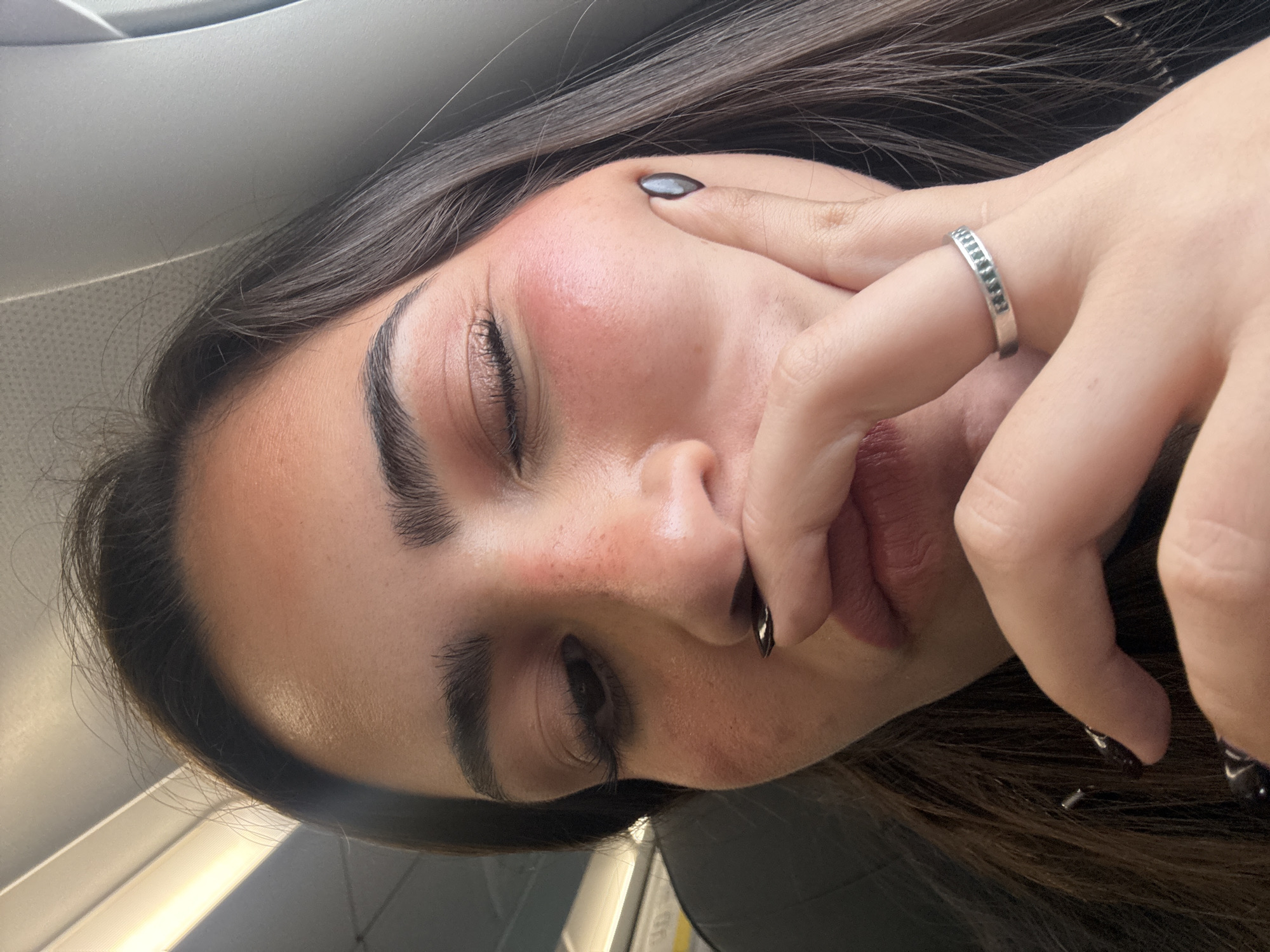A young woman with dark hair and dark nail polish is positioned in what appears to be the interior of a vehicle. Her face is turned to the side with her eyes mostly closed. She is touching her face with one hand, on which she is wearing a silver ring. The background shows elements of a car's interior. The location in the city of San Antonio, United States of America is not evident in the image.
