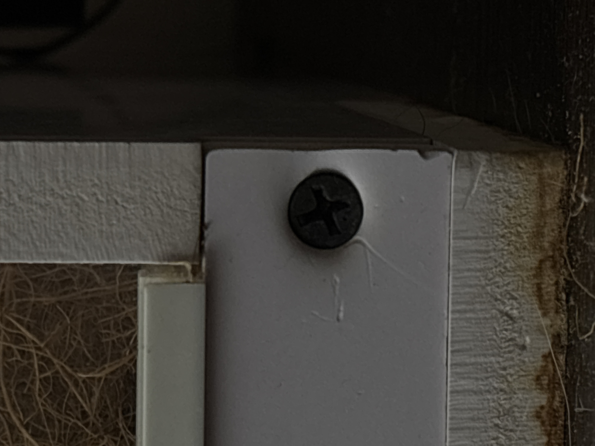 The image is a close-up shot of a white, textured surface with a black screw visible on it. The screw appears to be a Phillips head screw and is partially embedded in the surface. The surface itself is slightly dirty at the bottom, with some brown staining. To the left of the screw, there are some faint white scratch marks on the surface. The background is dark and out of focus. The image does not contain any elements that would suggest a location in Saint Petersburg, Russia.