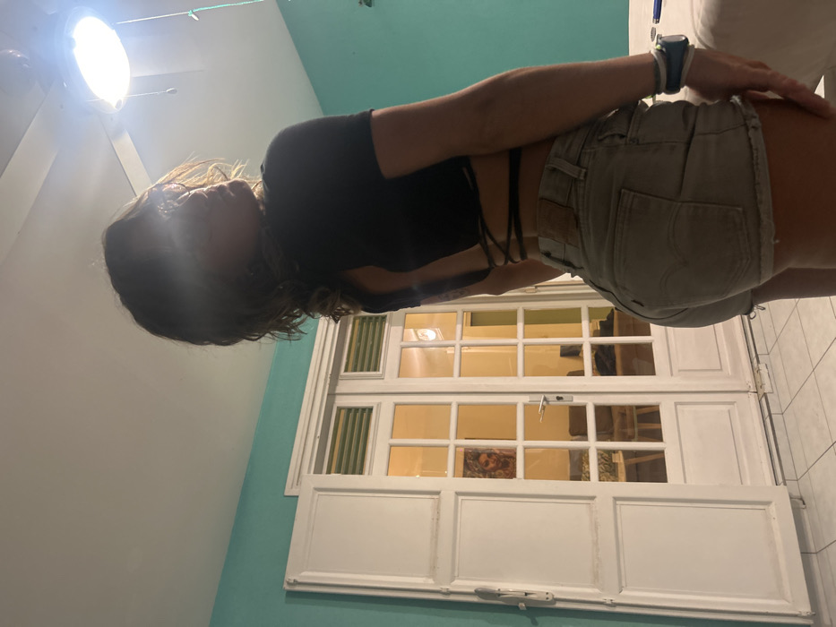 An interior scene in Sainte-Anne, France, depicts a female subject from a rear, slightly left-side perspective. The subject has wavy, light-brown hair and is wearing a black, short-sleeved crop top with an open, criss-cross laced back, paired with light-colored high-waisted denim shorts featuring a Levi's brand patch on the waistband. Her left wrist displays multiple wristbands. Her face is obscured by a strong light flare emanating from a white ceiling fan with an integrated light fixture, visible in the upper right. The room features teal-colored walls and a light-colored tiled floor. In the center background, white double French doors with glass panes open into an adjacent, brightly lit interior space where a sofa is partially visible. A distinct reflection of a woman with blonde, curly hair is present in the upper-left pane of the French doors. To the left of the doors, a partially open white shutter reveals horizontal window slats. A white, possibly cushioned, surface is visible on the far right foreground.