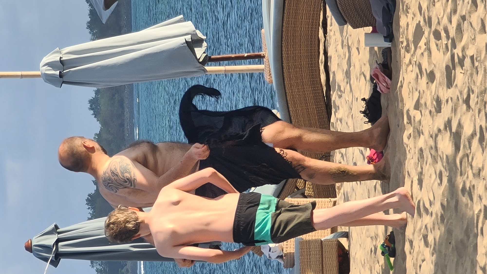 The image depicts a beach scene, likely in Denpasar, Indonesia, with two individuals standing on the sand near the ocean. The person in the foreground, a young boy, is facing away from the camera and is wearing swim trunks with horizontal bands of black and teal. Beside him stands a man, also facing away, with a tattoo on his left shoulder blade. He is wearing a dark sarong wrapped around his waist and appears to be in the process of adjusting it. In the background, there is the blue ocean, a line of trees, and two large beach umbrellas. The sand is light-colored and textured, with a few items like shoes and a white cylindrical object visible on the ground. The lighting suggests it is daytime, possibly late afternoon or early morning, with soft sunlight illuminating the scene.