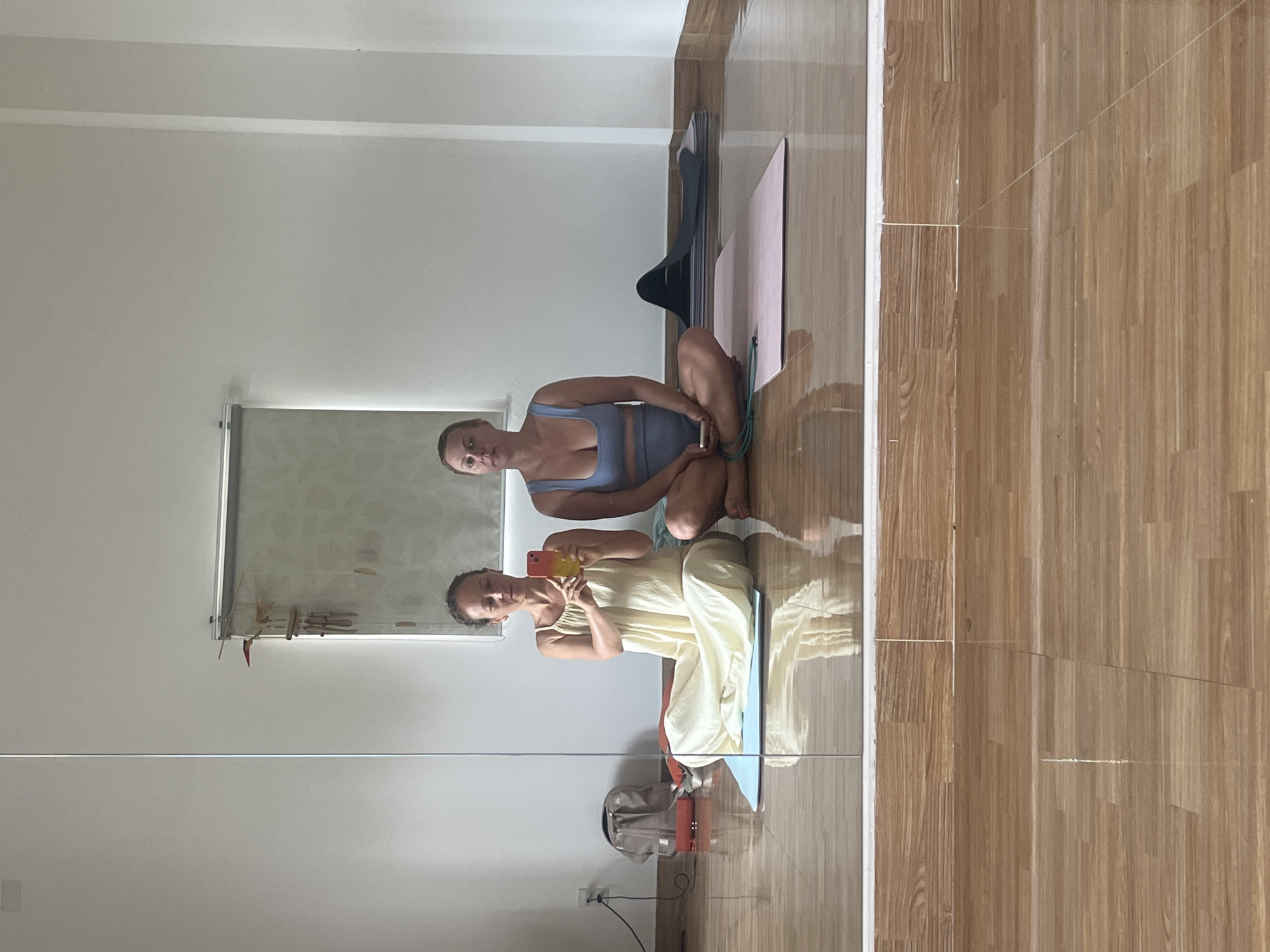 Two women are seated on yoga mats on a wooden floor in what appears to be a yoga studio. The woman on the left is wearing a long, light-colored dress and is holding a smartphone to take a selfie. The woman on the right is wearing a blue yoga set and is looking directly at the camera. Several yoga mats and what appears to be a resistance band are visible on the floor. In the background, there is a large mirror reflecting the scene, a window with blinds, and a wall-mounted electrical outlet. A backpack and a red object are also visible in the reflection. The overall lighting suggests it is daytime. The scene is located in Đà Nẵng, Vietnam.
