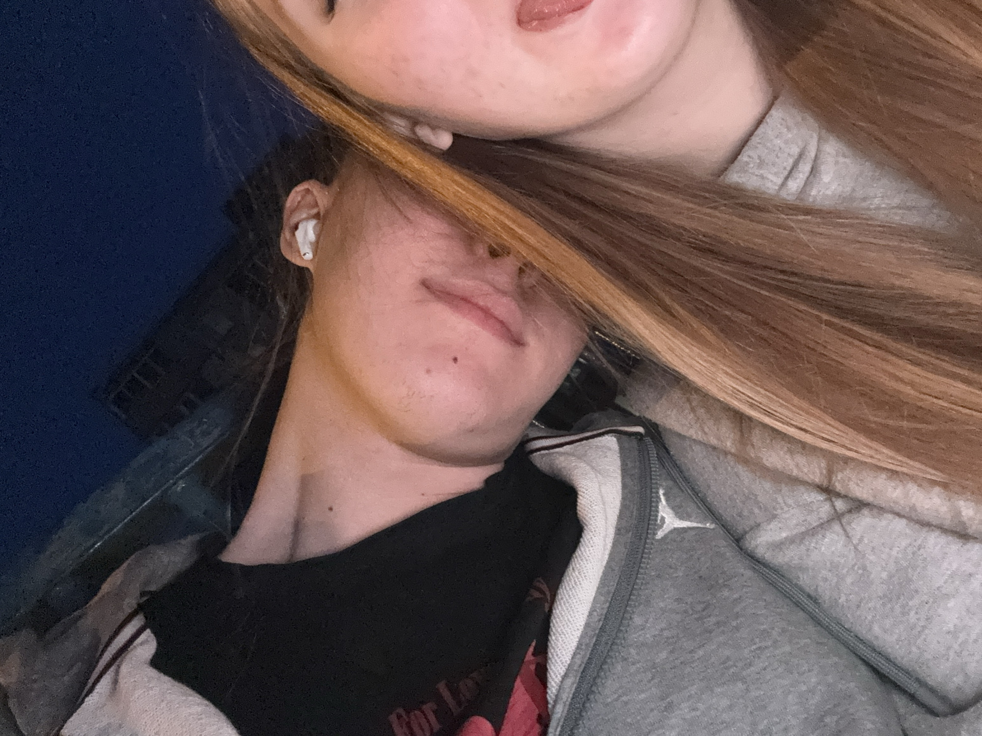 Two individuals are captured in a close, tilted shot, likely a selfie. One individual, positioned lower in the frame, is wearing a black t-shirt under a grey zippered jacket, with fair skin, a mole, and a visible earbud in their ear. The other individual, partially visible at the top, has fair skin with some blemishes and long, light brown hair. The background shows a dark blue sky and the vague outline of a building. It is unclear if the location is Perm, Russia.