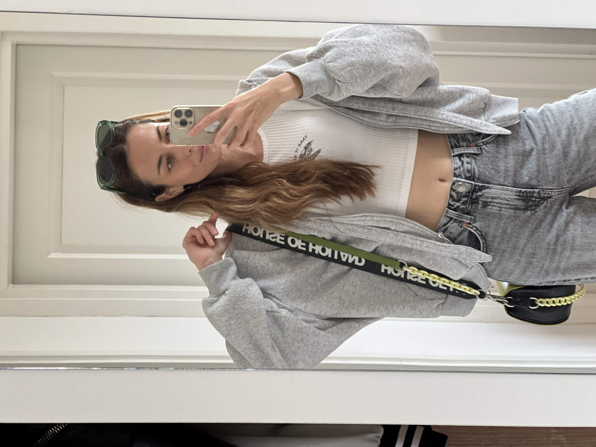 A woman takes a selfie in a mirror. She wears sunglasses perched on her head, a white crop top, a light grey zip-up hoodie, and light grey jeans. She holds a gold-colored smartphone in her right hand. A black purse with a yellow chain is slung over her shoulder, the strap bearing the words "HOUSE OF HOLLAND". The mirror reflects a white panelled door. The location is likely indoors, possibly a residence or a changing room, but there are no discernible details to confirm it to be Bar, Montenegro.