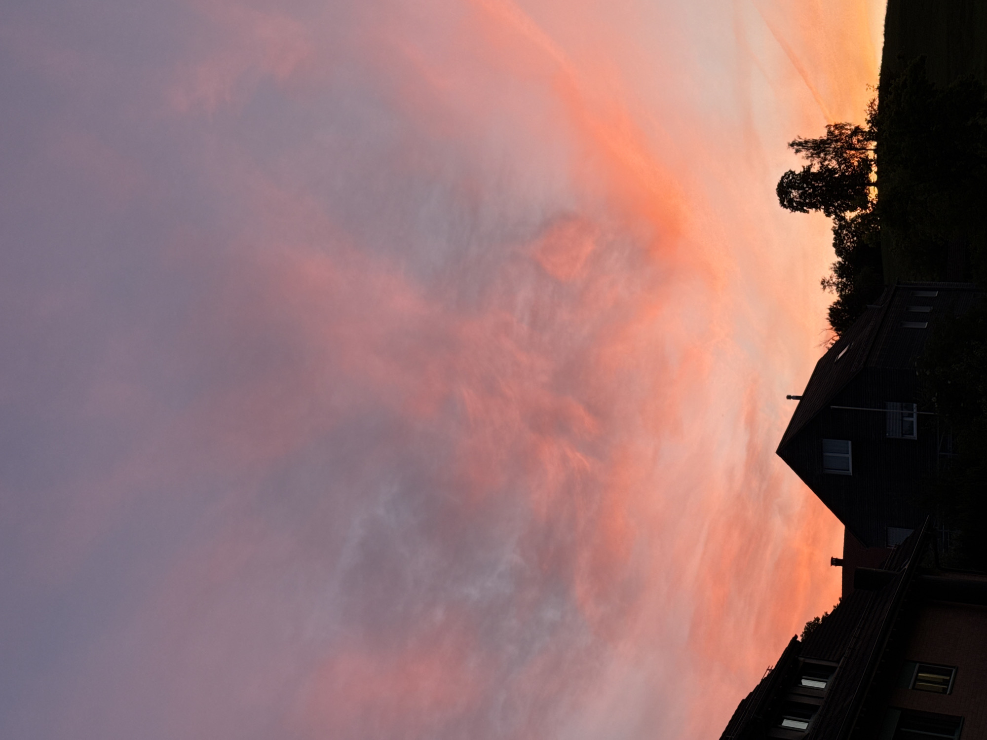 This is a vertical, eye-level, medium shot of a sunset in Lufingen, Switzerland. The sky is a vibrant gradient of pink, orange, and purple, with wispy clouds scattered across it. In the foreground, the silhouetted rooftops of several houses are visible. A large tree stands on a hill to the right of the frame. The overall impression is one of peacefulness and natural beauty.