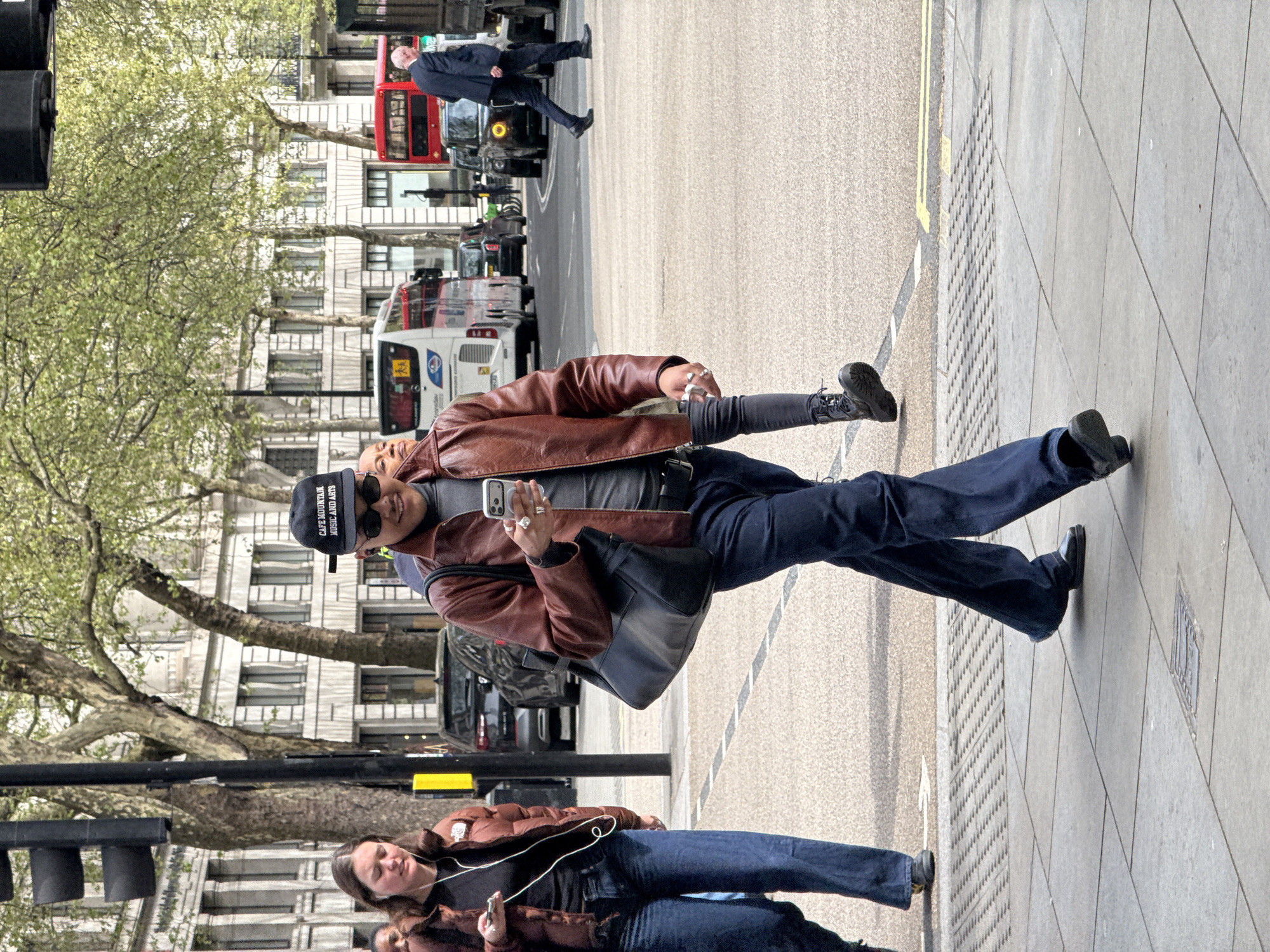 A street scene in London, United Kingdom, shows a man wearing a brown leather jacket, dark blue jeans, and black shoes walking on the sidewalk. He is holding a smartphone in his right hand and wearing a black baseball cap with white lettering. A shoulder bag is slung across his body. Behind him, a woman in a similar jacket and jeans walks with headphones. In the background, London buses, cars, and buildings are visible. A double-decker bus is prominent. A man in a dark suit walks across the street in the background. Trees line the street. The streetlights have not been activated.