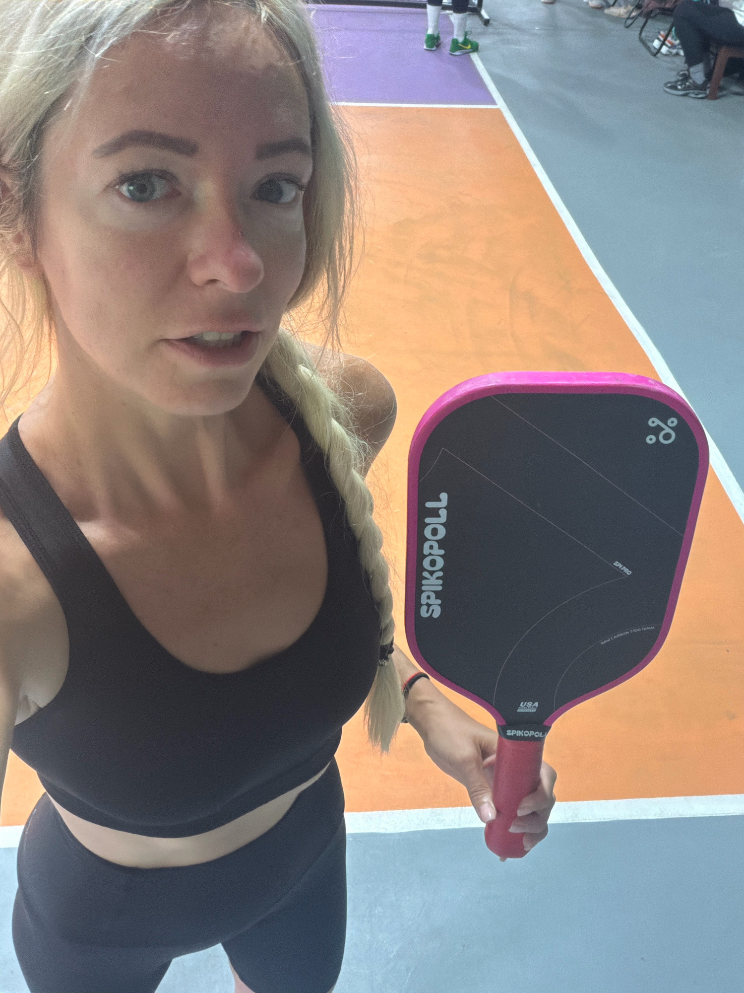 A woman is posing for a selfie on a pickleball court. She is wearing a black sports bra and black athletic shorts, and she has blonde hair in a braid. She is holding a pink and black pickleball paddle. The court has orange and purple sections, with grey flooring. In the background, other people are visible, some seated and some standing. The scene appears to be indoors, as indicated by the flooring and lighting. There is no information in the image to suggest the location is Da Lat, Vietnam.