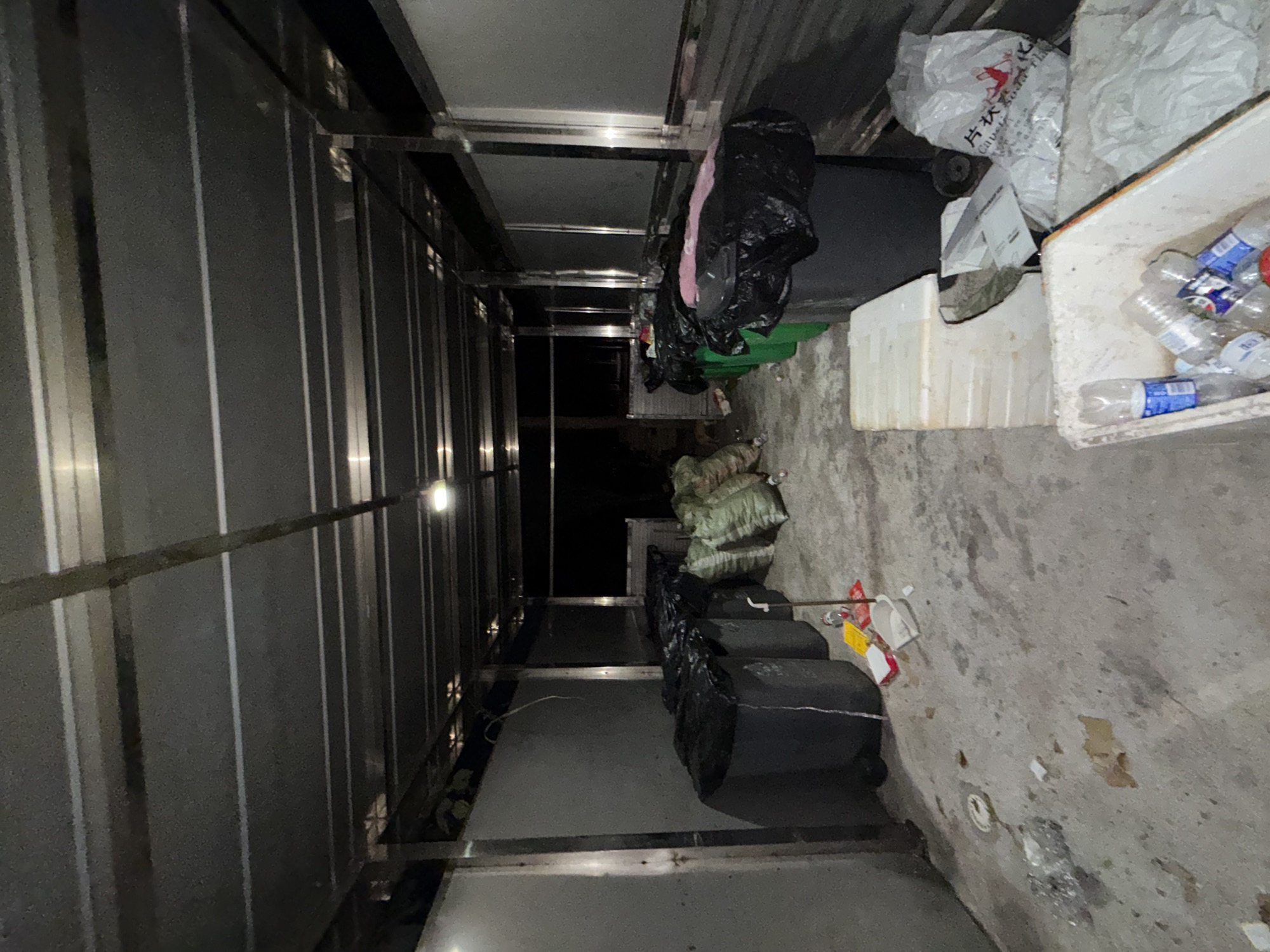 The image depicts an outdoor covered area, likely a waste disposal or collection point, with a concrete floor and a metal roof supported by metal beams. The area is lined with various trash bins and bags. On the left side, several large, dark trash bins are visible, some covered with black plastic. To the right of these, there are several large, green mesh bags filled with what appears to be refuse, possibly recyclables. Further to the right, more large bins are present, also covered with black plastic. In the foreground, there are some scattered items on the concrete floor, including a white styrofoam container filled with plastic bottles and other debris, as well as some discarded packaging. The scene is dimly lit, suggesting it may be nighttime or an overcast day. The overall impression is one of a functional, albeit somewhat unkempt, waste management area. The specific location is a waste collection point in the city of Jiangjin, China.
