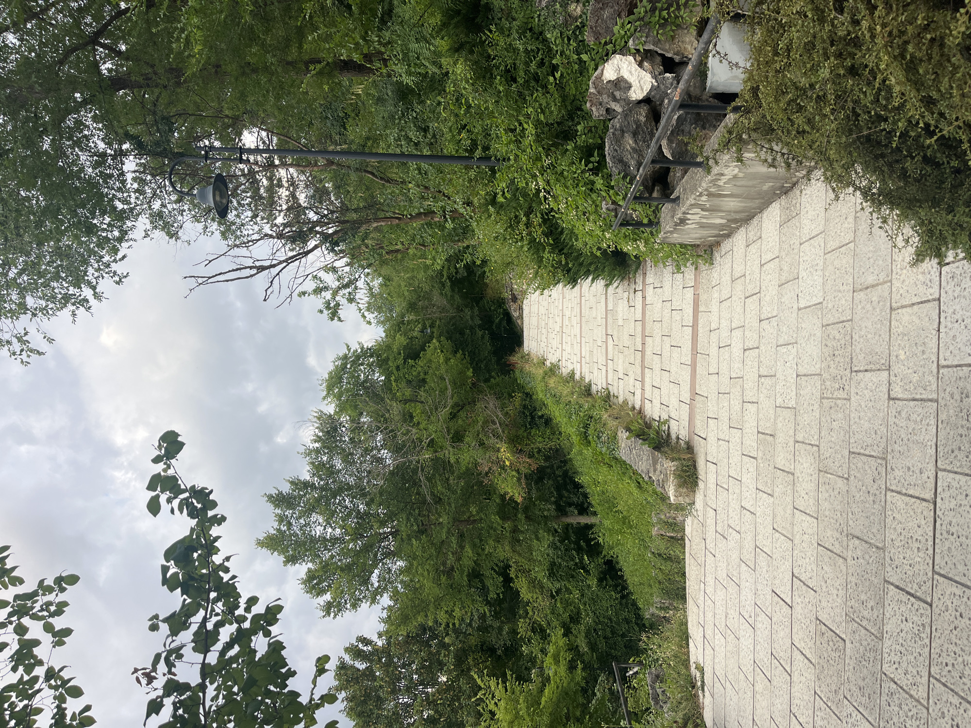 An outdoor, slightly overcast daytime shot shows a wide, paved stone walkway leading uphill through a dense, green forest. The walkway, composed of large, rectangular light-colored stone tiles, is bordered on the left by a low stone wall and lush green vegetation, and on the right by a black metal railing, a concrete retaining wall, and a cluster of large rocks. A single, dark lamppost with an old-fashioned lantern is visible on the right side of the walkway, surrounded by trees and foliage. The trees form a thick canopy overhead, with branches and leaves visible in the upper portion of the frame. The overall impression is one of a natural, well-maintained pathway within a park or a wooded area. This scene is located at the Echo Park in Dobrich, Bulgaria.