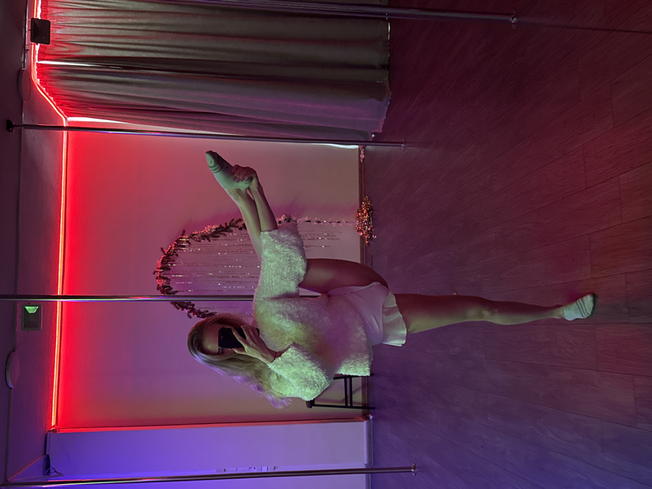 A full-shot, rotated image depicts a person performing a full split on a wooden floor in what appears to be a pole dancing or fitness studio. The person, with blonde hair, is lying horizontally on the floor with one leg extended forward and the other backward, demonstrating advanced flexibility. She is wearing a white, fluffy long-sleeved top, white shorts, and light-colored socks, and is holding a smartphone up to capture a selfie.
The indoor setting features a dark wooden floor and walls illuminated by vibrant LED strip lights – a bright red strip runs vertically along the left wall, and a purple/blue strip is visible along the bottom. Two metal poles, commonly used in pole dancing, frame the scene. In the background, a decorative element resembling a garland with shimmering, tinsel-like strands and small lights adds a festive touch. The overall lighting suggests an evening or nighttime setting. The person's face is largely obscured by the phone. No text is visible in the image.