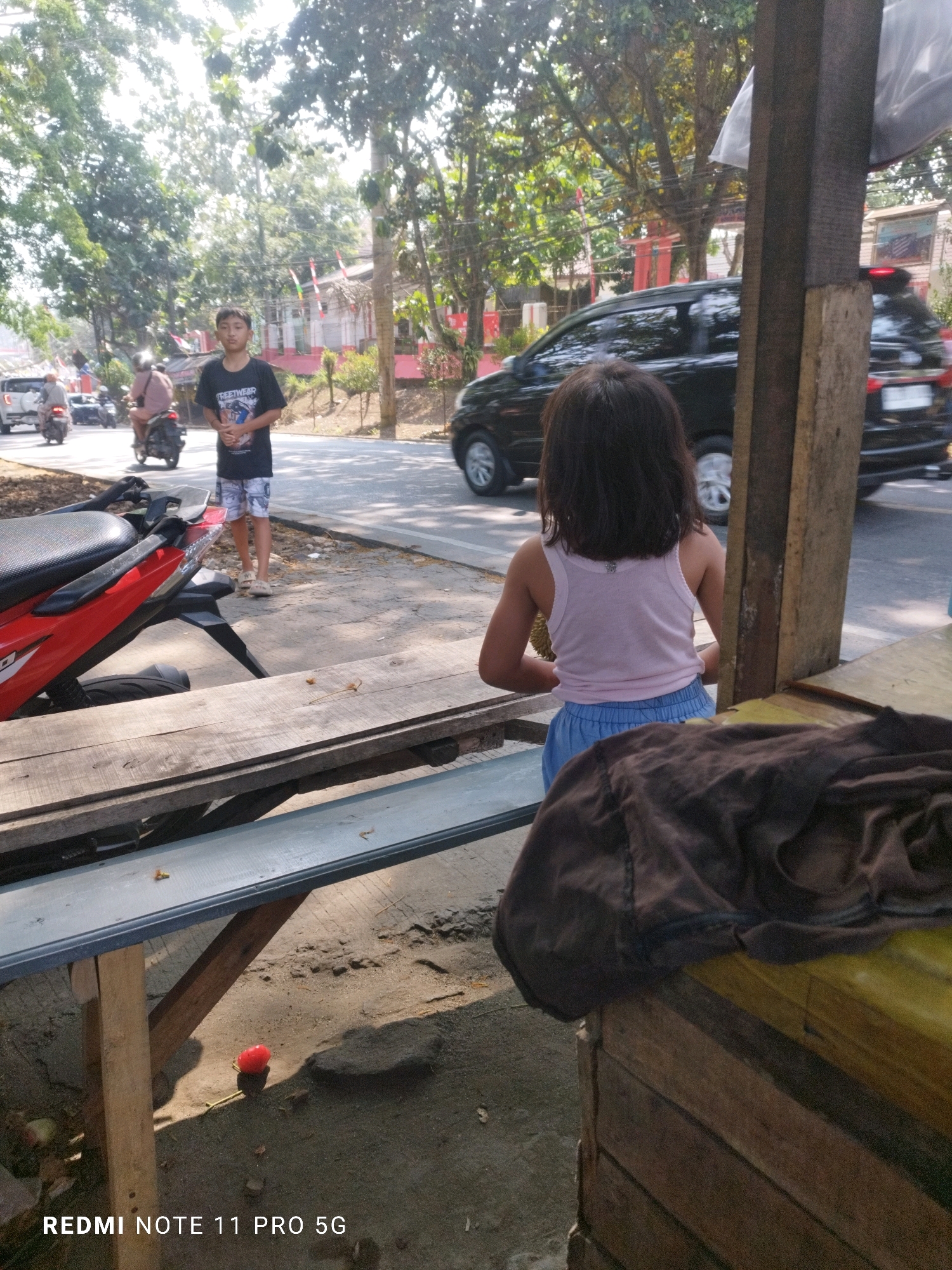 The image captures a street scene in Saruni, Indonesia, bathed in bright sunlight. In the foreground, a young girl with dark hair faces away from the camera, wearing a pink tank top and blue shorts, sitting at a wooden table. To her left, a red motorcycle is parked. Behind the girl, a young boy in a black t-shirt and patterned shorts stands on the side of a paved road. The road is busy with traffic, including cars and motorcycles. Trees line the road, and buildings with red accents are visible in the background. The overall atmosphere suggests a typical day in a bustling Indonesian town.