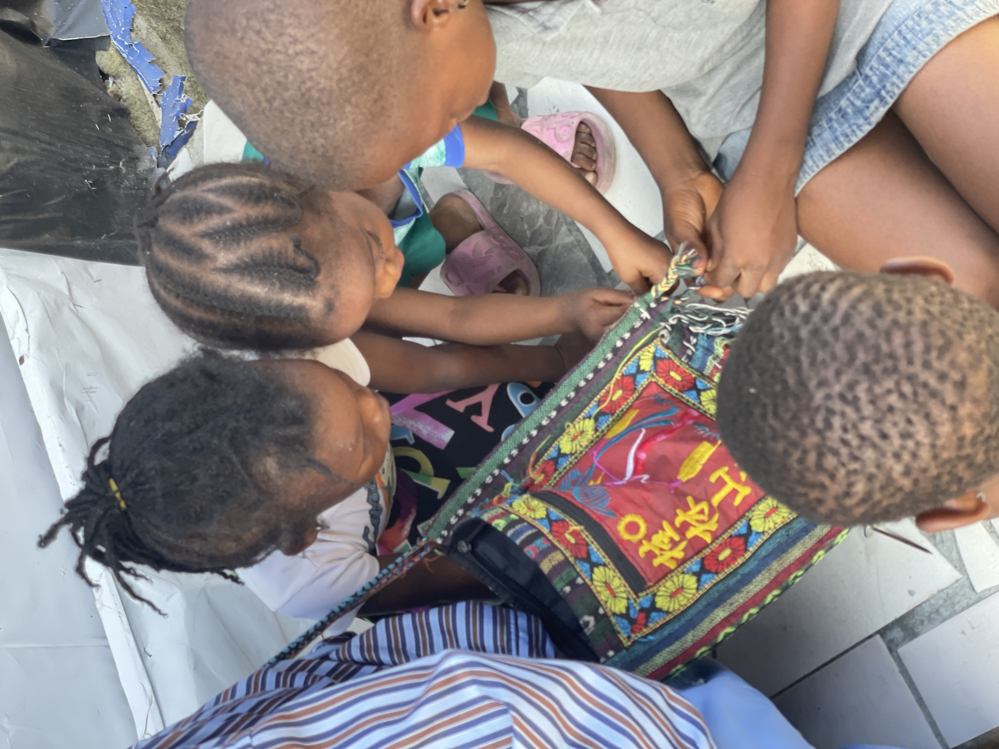 In Iranla, Nigeria, four young Black individuals are gathered around a vibrantly decorated bag, their attention focused on its contents and construction. One person, wearing a blue and white striped shirt, is partially visible on the left. Two younger children, one with dreadlocks and a yellow tie, the other with braided hair, are positioned between the first person and the bag. A third child, wearing a grey t-shirt and denim shorts, is on the right, intently working on the bag with the other two children. The bag itself is adorned with colorful embroidered patterns, featuring flowers and what appear to be yellow Chinese characters. The ground beneath them is a tiled surface, and a white tarp is visible in the background, suggesting an outdoor or makeshift setting. The children's actions indicate engagement in a shared activity, possibly repairing or embellishing the bag.
