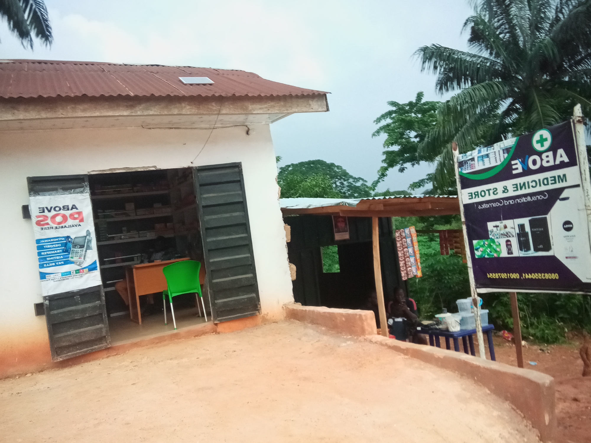 This is a medium shot, taken outdoors in what appears to be a rural or semi-urban area in Oko Town, Nigeria. The foreground is dominated by a reddish-brown dirt path. To the left, a white building with a corrugated tin roof, painted light yellow underneath, is partially visible. The building has an open doorway with dark metal shutters partially pulled back, revealing shelves stocked with various items inside. A poster is attached to the left wall outside the doorway, with text that appears to be a mix of English and possibly local language, featuring an image of a mobile phone. To the right of the building entrance, a bright green plastic chair with a metal frame is positioned near a wooden desk inside.
A dark wooden structure with a corrugated tin roof is attached to the right of the white building, providing shade. Underneath this structure, a person, possibly a man, is seated behind a small table, appearing to be tending to items. Several plastic containers are visible on the table.
On the far right of the image, a large vertical sign is erected. The sign is dark purple and white, featuring a green cross emblem at the top, followed by text that reads "ABOVE MEDICINE & STORE". The sign also displays images of various products and a phone number. The background consists of lush green trees and vegetation, with a palm tree prominent on the far right. The sky is overcast and appears to be a pale grey. The overall impression is of a small, open-air shop or convenience store in a tropical environment.