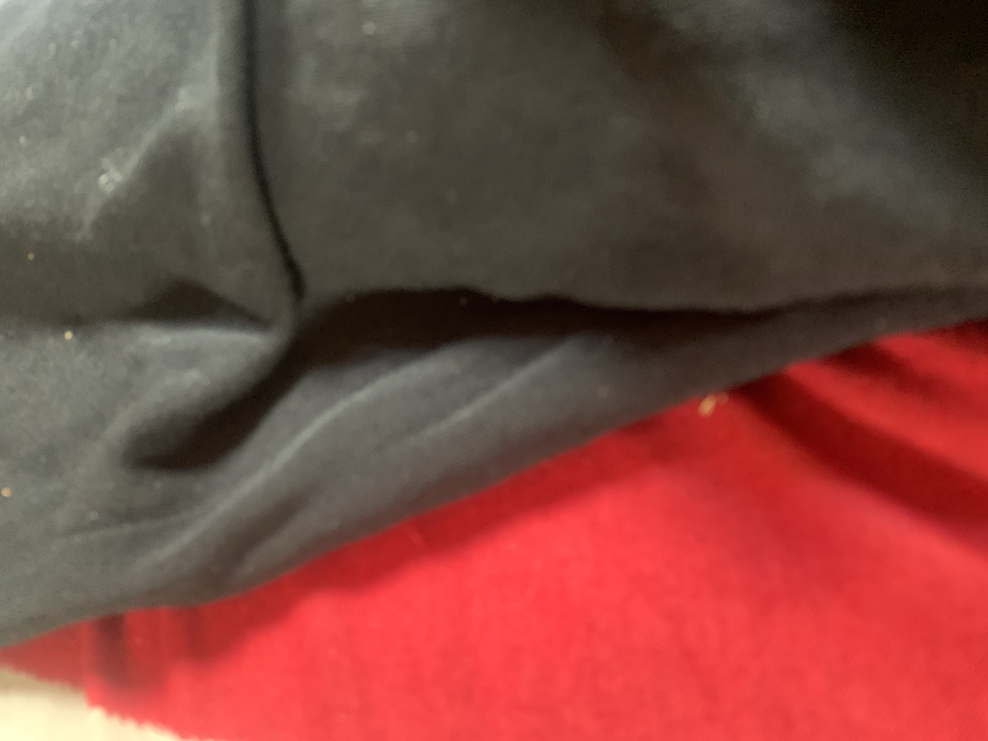 The image shows a close-up view of two pieces of fabric, one red and one dark gray, with a clear division between them. The fabrics are draped and folded, creating shadows and highlights that reveal their texture. The red fabric appears to have a slightly rougher texture, while the dark gray fabric seems smoother and possibly made of a different material. There are a few small, light-colored specks visible on the dark gray fabric, which could be dust or lint. The lighting suggests an indoor setting. There is no identifiable information in the image that would place it in Jauro Yinu, Nigeria.
