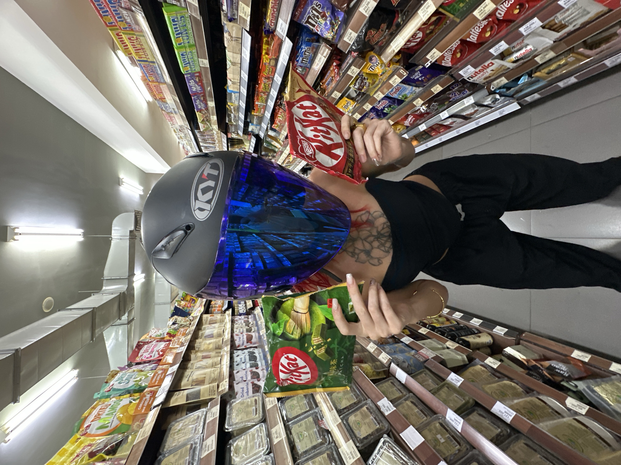 A person wearing a gray motorcycle helmet with a blue visor is standing in a supermarket aisle in North Kuta, Indonesia. The person is holding two packages of Kit Kat bars - one a traditional red wrapper, and the other in green with matcha flavoring. The shelves surrounding them are stocked with various candies, chocolates, and dried fruits.