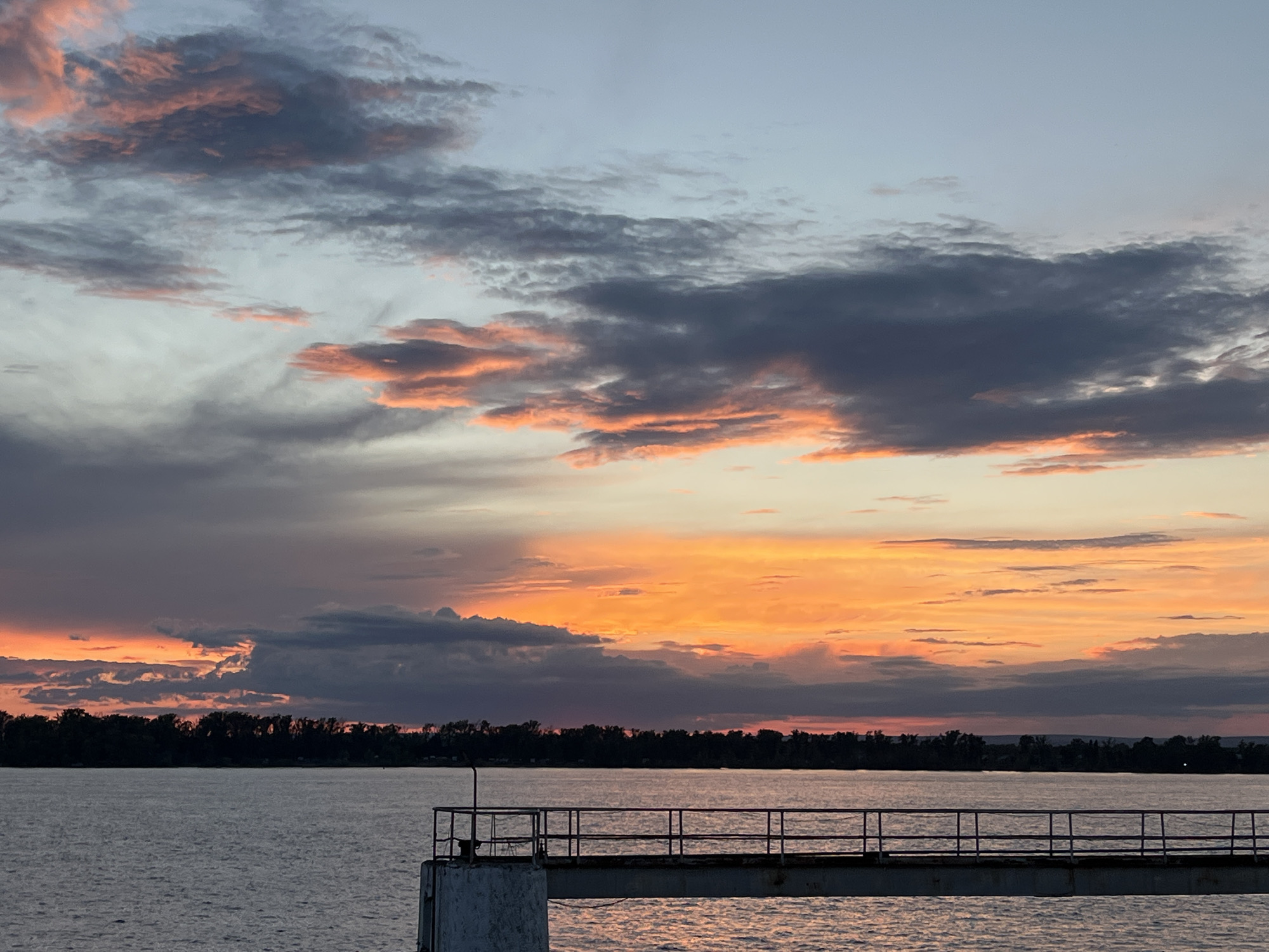 This media file depicts a serene sunset scene over a body of water, likely a river or lake, near Novokuybyshevsk, Russia. The sky is ablaze with vibrant hues of orange and pink, transitioning into softer tones of blue and gray as it stretches towards the horizon. Wispy clouds are scattered across the sky, catching the last rays of sunlight and creating a dramatic and colorful display.
In the foreground, a portion of a pier or dock juts out into the water. It is constructed of metal railings and a concrete base, appearing to be sturdy but weathered. The water itself is calm, with a gentle ripple effect reflecting the colors of the sunset. A dark silhouette of a tree line or forest borders the far bank of the water, providing a sense of depth and natural beauty to the landscape.
The overall mood of the image is peaceful and contemplative. The time of day is clearly sunset, as indicated by the warm, vibrant colors in the sky. The weather appears clear and calm, with no visible signs of rain or wind. The location, Novokuybyshevsk, Russia, suggests a potentially industrial or rural setting, though the natural beauty of the sunset dominates the scene. There is no visible text in the image.