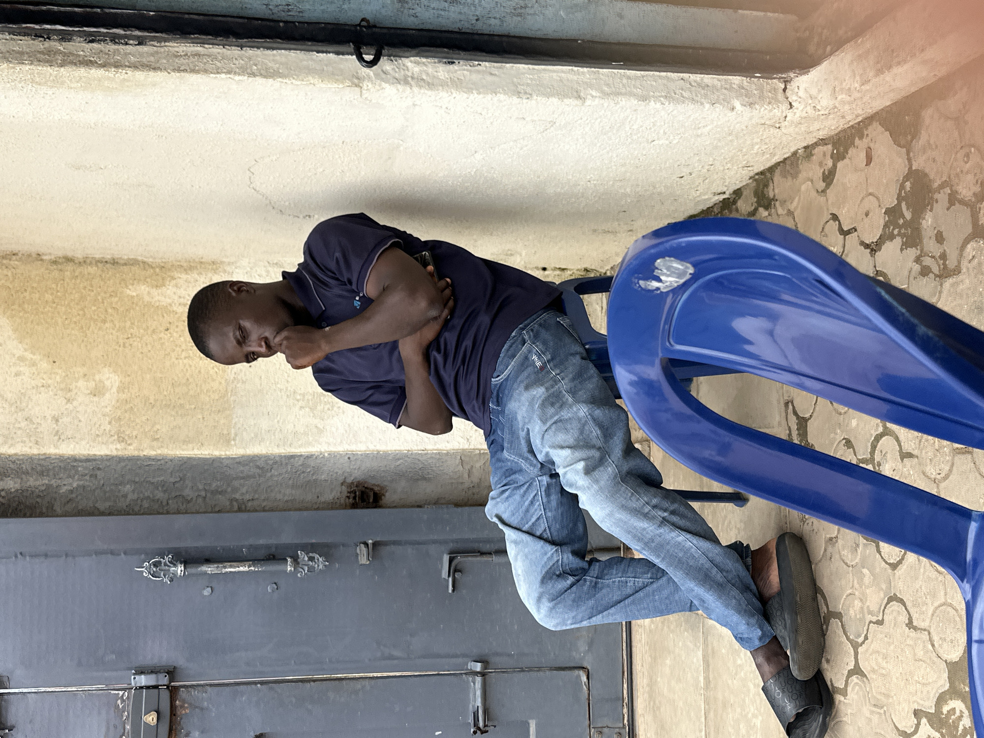 A young Black male is seated on a blue plastic chair next to a light-colored concrete wall in Narayi, Nigeria. He is wearing a dark blue polo shirt and light-wash denim jeans. His left arm is crossed over his chest, and his right hand is touching his chin, suggesting a pensive or thoughtful mood. His legs are crossed at the knees, and he is wearing dark, open-toed sandals. To his left, a dark gray metal door with decorative handles and visible locking mechanisms is present. In the foreground, another blue plastic chair is partially visible. The ground is paved with interlocking concrete blocks, some of which appear wet. The wall behind him has a textured surface with some water stains.
