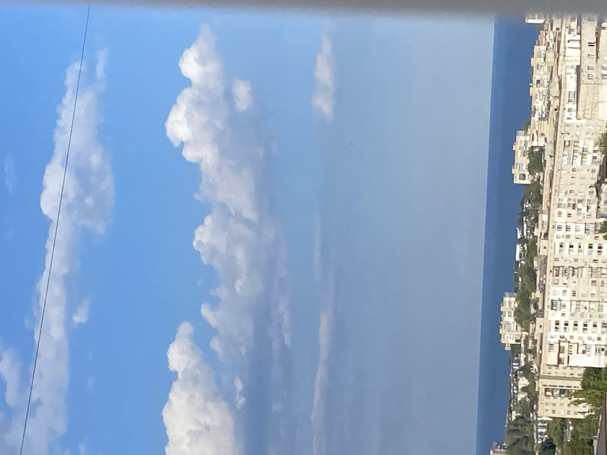 The image depicts a coastal city with residential buildings situated along the shoreline. The sky is blue with scattered white clouds, and a thin, dark line, possibly a wire, stretches horizontally across the upper portion of the frame. Below the sky, a body of water, likely the sea, forms a distinct horizontal band. In the foreground and midground, numerous multi-story residential buildings with visible windows and balconies are densely packed together. Green trees and vegetation are interspersed among the buildings. The scene appears to be an urban landscape in Molodizhne, Ukraine, on a clear day.