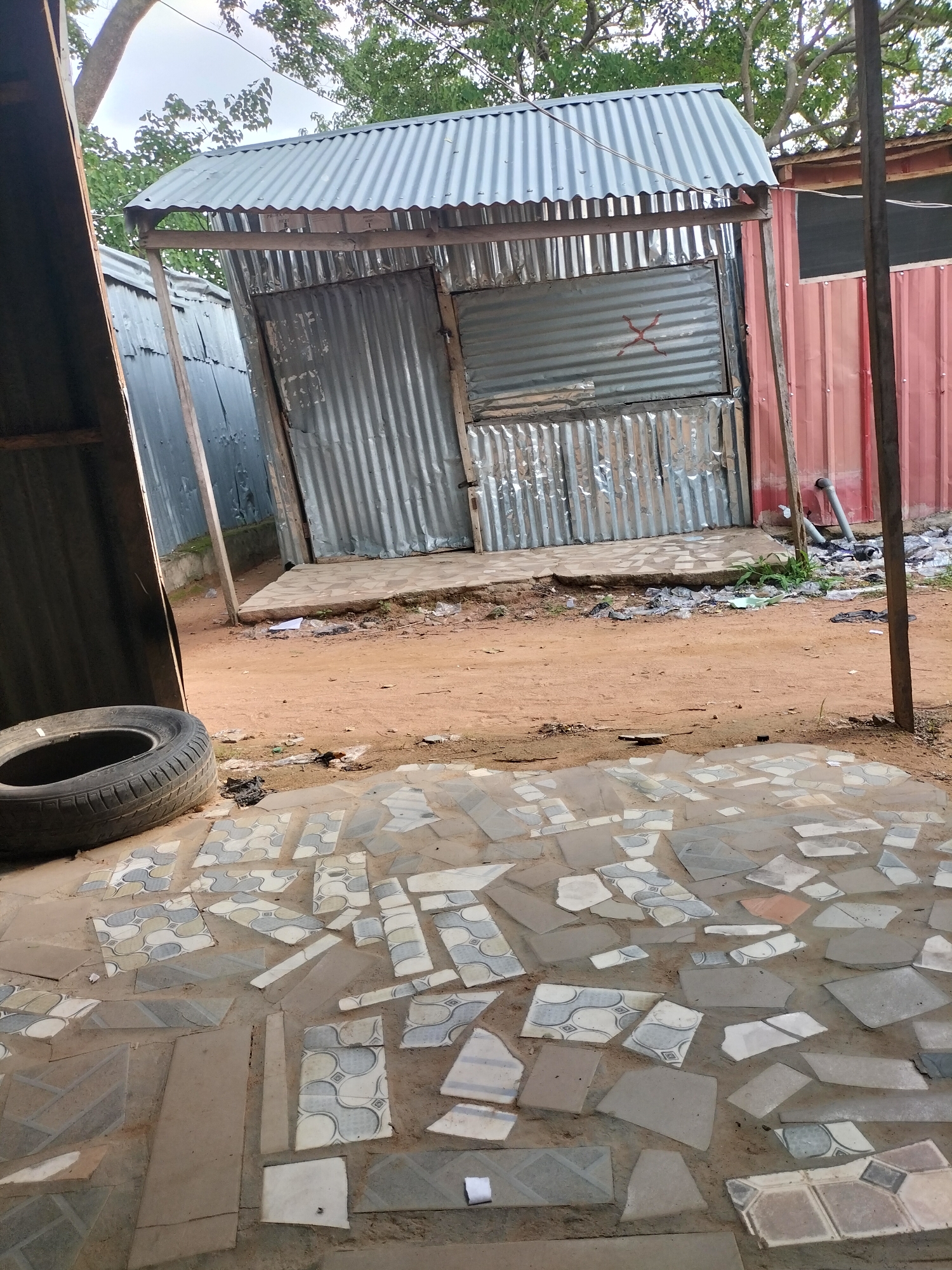 The image depicts an outdoor scene in Jalingo, Nigeria, with a focus on a makeshift structure constructed from corrugated metal. The structure appears to be a small stall or shop, with a sloped roof made of the same material. The front of the stall is open, revealing a rough, dirt floor. The walls are also constructed from corrugated metal sheets, some of which are dented or damaged. A section of the wall has what looks like a red "X" mark painted on it.
To the left of the stall, part of another corrugated metal structure is visible, leaning inwards. In the foreground, a weathered tire lies on the ground. The ground itself is a mixture of red dirt and scattered, broken tiles, creating a mosaic-like pattern in places. There is some litter scattered around the base of the stall.
In the background, lush green trees with visible branches suggest an outdoor setting with natural foliage. The sky is overcast, indicating a cloudy day. The overall impression is one of a developing or informal settlement, where structures are often built with readily available materials. There is no visible activity or people in the image. The time of day is indeterminate due to the overcast conditions, but it appears to be daytime.