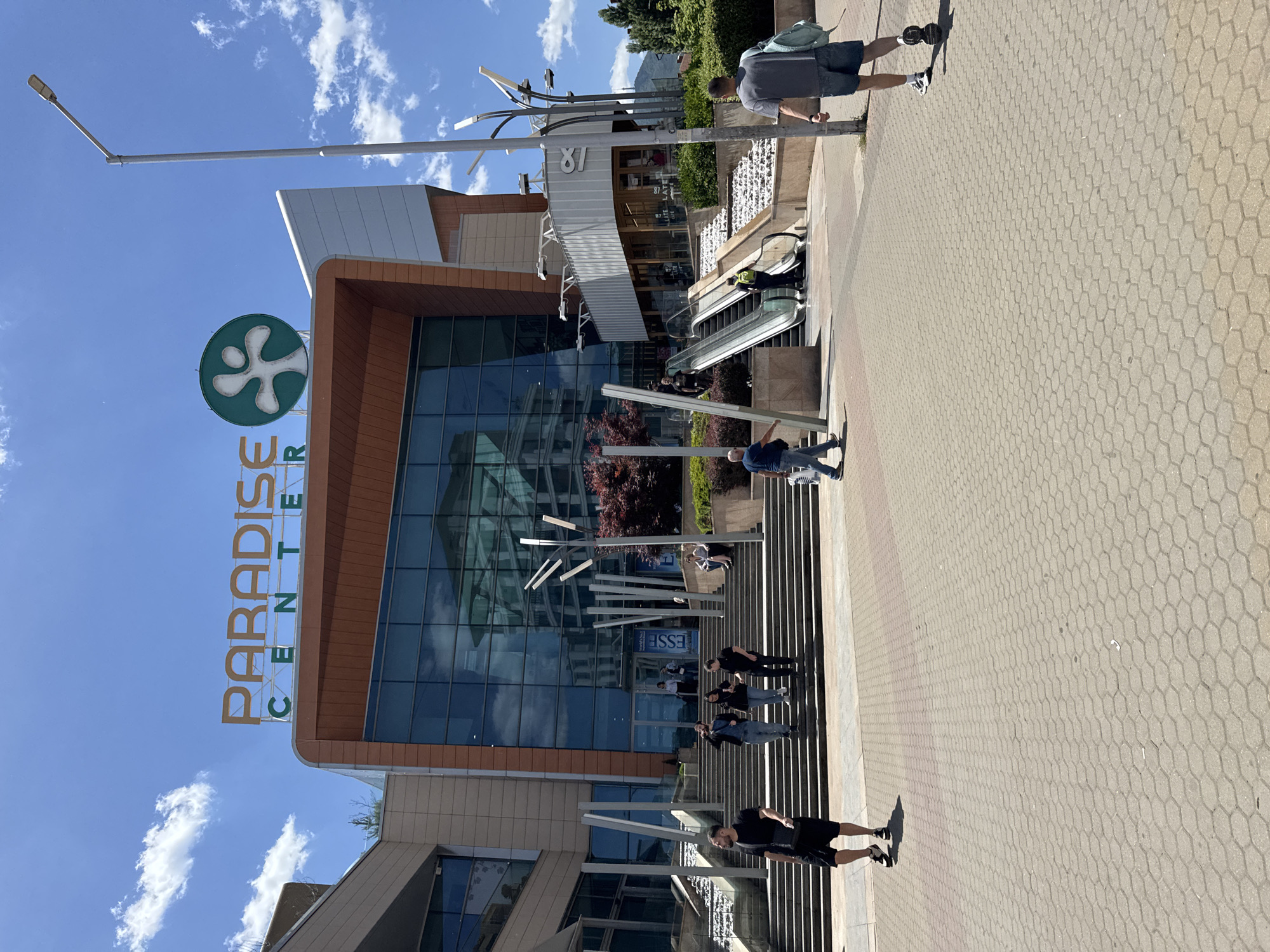 The image depicts the Paradise Center in Sofia, Bulgaria. The building has a modern design with a large glass facade and brown wood paneling. The Paradise Center logo is visible on the left side of the building. Several people are walking on the stairs leading to the entrance. There is an escalator on the right side of the building. The foreground shows a paved walkway with a geometric pattern. The sky is blue with scattered clouds. A tall lamppost is visible on the left side of the image.