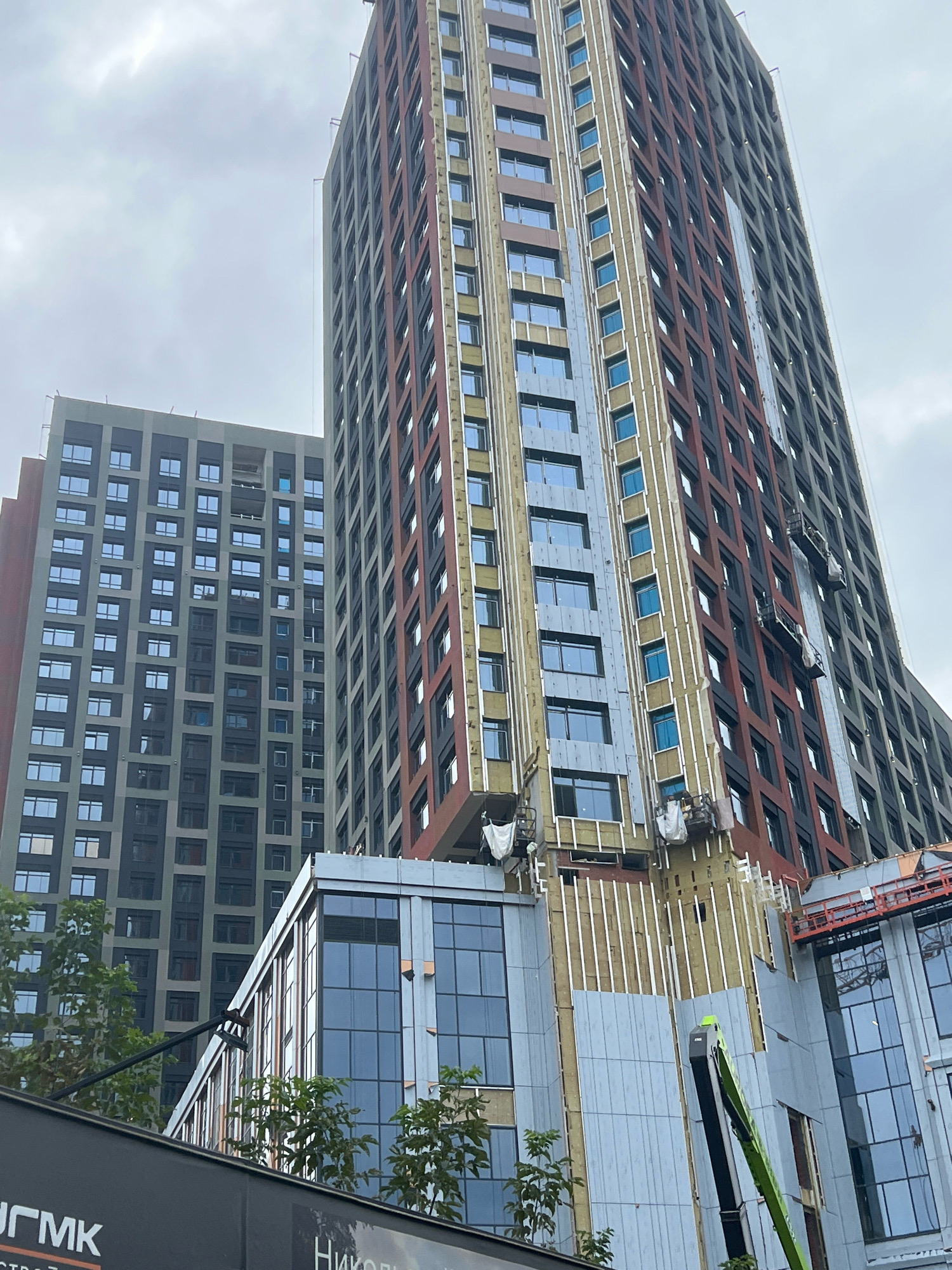 The image shows the construction of high-rise buildings in Yekaterinburg, Russia. The main focus is on two tall buildings with distinctive facades. The building on the right features a vertical pattern of reddish-brown and dark gray panels, interspersed with lighter colored sections and visible insulation. Two suspended work platforms with workers are attached to the side of this building, indicating ongoing construction or facade work. The building on the left is a uniform gray with a grid of windows. In the foreground, there is a lower, modern building with large glass windows, also under construction, with scaffolding visible. Green trees are growing in front of the lower building. The sky is overcast with clouds. Partially visible signs in Russian are present at the bottom of the image.