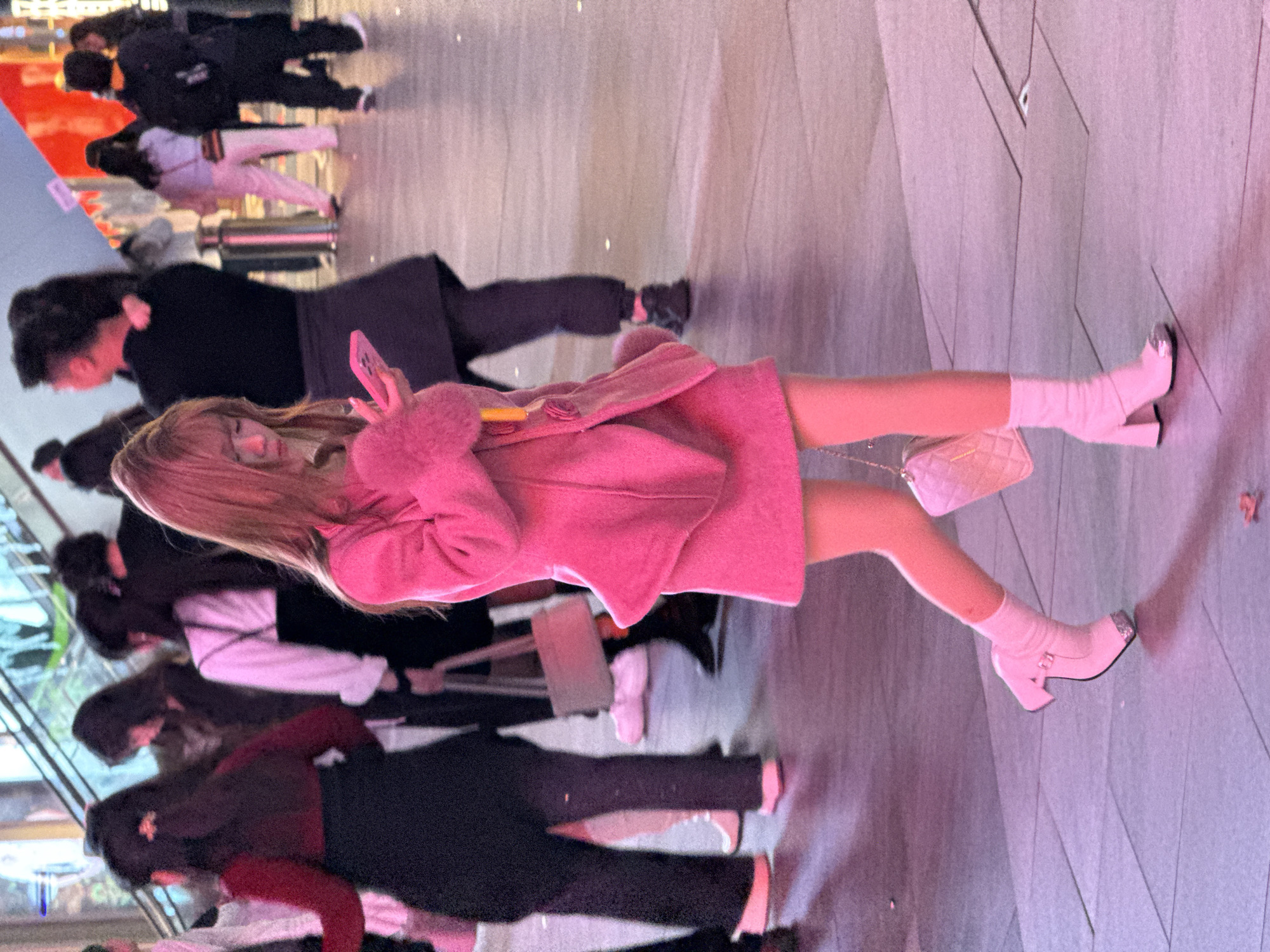 The image captures a bustling urban pedestrian scene in the city of Liangjiang, China, under artificial evening or nighttime lighting, which casts a dominant pinkish-purple hue across the environment.
The central subject is a young individual with long, light brown hair and bangs, walking from left to right while looking down at a pink smartphone held in both hands. They are dressed in a vibrant pink matching outfit: a structured coat with faux fur cuffs and a peplum hem, worn over a mini-skirt. A yellow detail is visible near the coat's lapel. Their lower legs are bare, complemented by white ruched ankle socks and pink ankle boots featuring chunky block heels and reflective silver toe caps. A quilted pink shoulder bag with a chain strap hangs from their left side.
In the background, numerous other individuals are visible, mostly blurred, indicating movement within a busy public space. To the left, a person in a red long-sleeve top and black skirt walks away from the viewer. Further back, another person is seen wearing a white long-sleeve top under a dark, possibly pinafore-style, dress, carrying a light-colored rectangular handbag. Other figures in the mid-ground and background are dressed in a mix of dark and lighter clothing, with one person observed in light pink pants. An escalator with glass side panels is partially visible on the left, with individuals ascending. The ground is a light-colored, paved surface, appearing to be composed of square panels, reflecting the ambient lighting, and contains minor scattered debris.