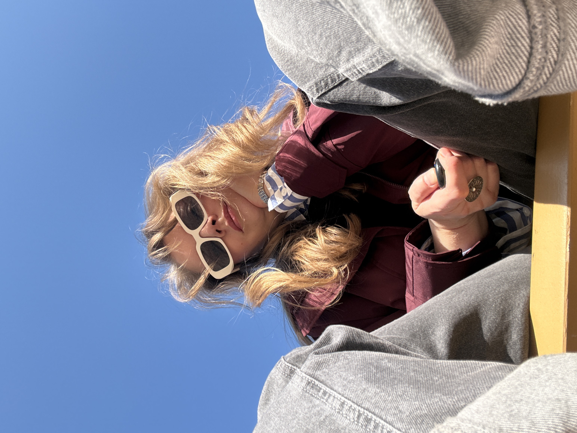 A low-angle shot looking up at a person wearing white-rimmed sunglasses, a maroon jacket over a blue and white striped shirt, and grey jeans. The person is wearing two rings on their right hand, one black and one silver. The background is a clear blue sky. There is a yellow object in the bottom right corner of the frame. The location is indeterminable.