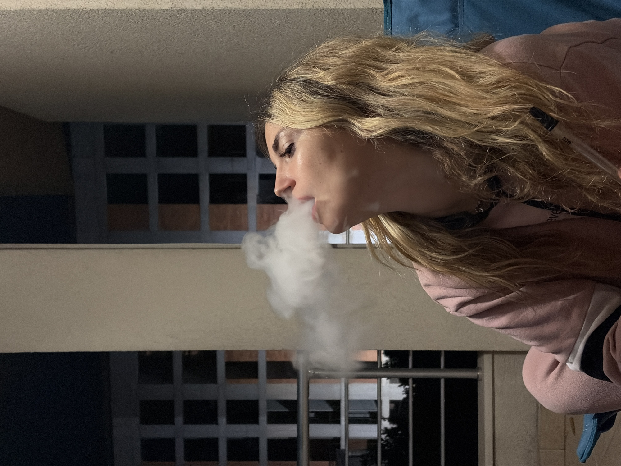 A woman is exhaling vapor, possibly from an e-cigarette or similar device. She has long blonde hair and is wearing a pink sweatshirt. The background consists of what appears to be the exterior of a building with multiple windows. The location is unspecified, however, the request mentioned the city of Budva, Montenegro.