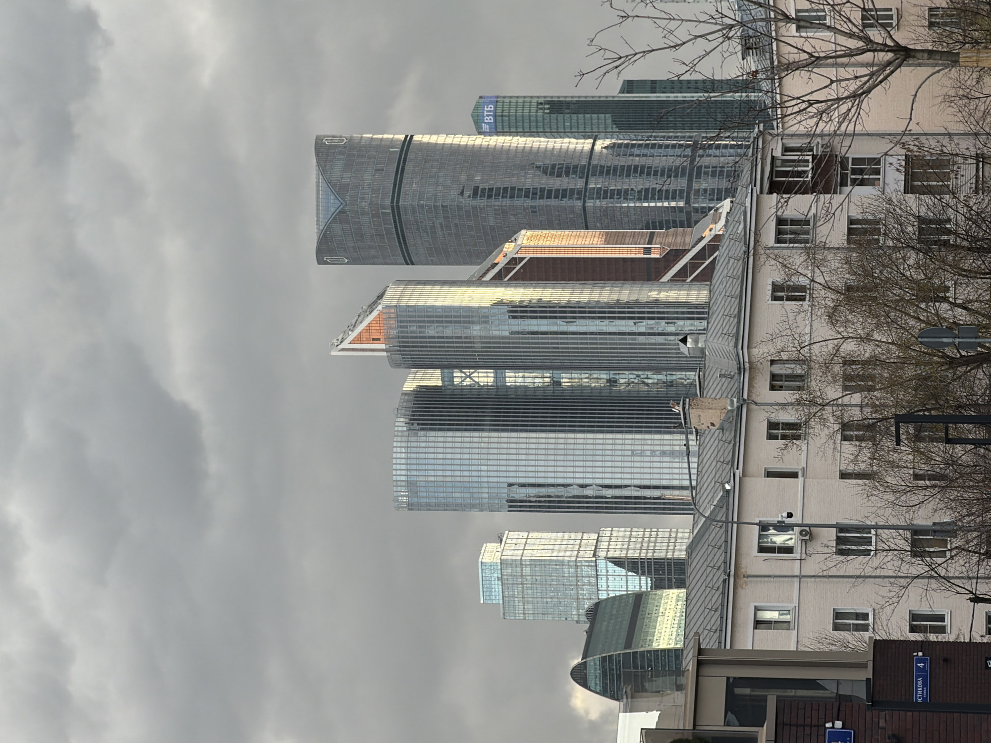 The image depicts a view in Moscow, Russia, juxtaposing modern skyscrapers with a more traditional residential building. The skyline is dominated by several high-rise buildings with reflective glass facades. One tower has a "ВТБ" logo at the top. An older, light-colored residential building with multiple windows is situated on the right side of the image. Leafless tree branches are visible in the foreground, partially obscuring the building. The sky is overcast, with grey clouds filling the upper portion of the frame. A street sign is visible in the lower right corner.