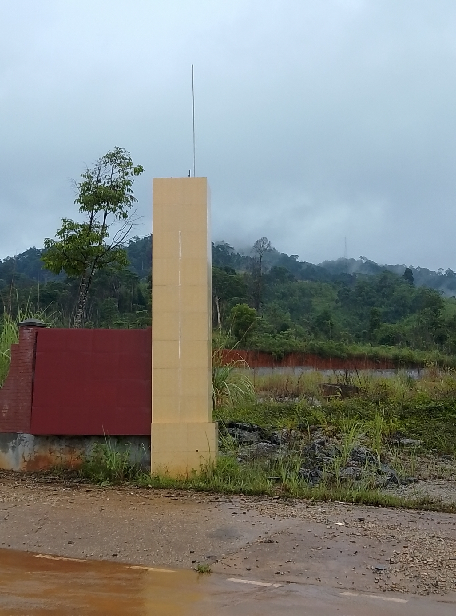 The image captures a rural or natural landscape under an overcast sky. In the foreground, a wet, unpaved road with visible puddles and gravel covers the bottom section of the frame, suggesting recent rainfall.
The central subject is a tall, rectangular column, light yellow or beige in hue, appearing to be constructed from panels or tiles. A slender, vertical antenna extends from the top of this column into the sky. To the immediate left of the column, a red or maroon wall structure, featuring visible brickwork on its left side, stands partially obscured by the column. Lush green vegetation, including tall grasses and smaller plants, grows around the bases of these structures and along the roadside.
The mid-ground and background are dominated by dense, verdant forests covering rolling hills and mountains. The upper portions of these mountains are partially shrouded in mist or low-hanging clouds, indicating high humidity or a cloudy weather condition. A faint, distant tower-like structure is discernible on one of the misty mountain ridges. The sky overhead is uniformly light gray, characteristic of an overcast day.
No people are visible in the scene, and there is no discernible text. The overall atmosphere is tranquil and natural, reflecting a wet, cloudy day in what appears to be a forested, mountainous region.