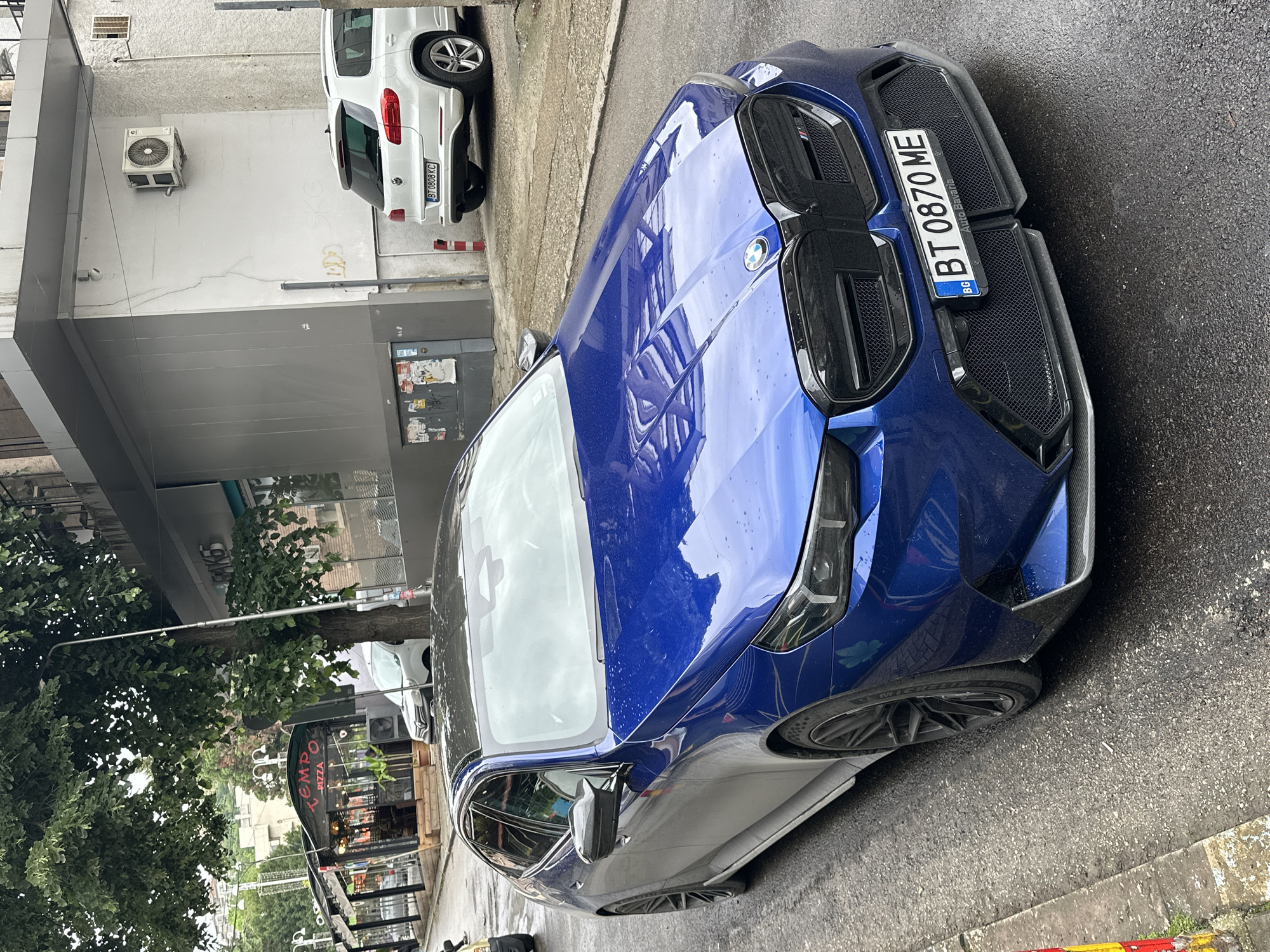 The image depicts a blue BMW parked on a street, likely in Veliko Tarnovo, Bulgaria, based on the license plate “BT 0870 ME.” The car is positioned at an angle, showcasing its front end, with water droplets visible on its surface. The reflections on the car's hood indicate surrounding buildings and trees. In the background, a white vehicle is partially visible, parked alongside a building with an air conditioning unit. Street-side establishments, including “Tempo Pizza,” can be seen with signage obscured by trees. The road surface appears to be wet. No people are discernible in the immediate foreground.
