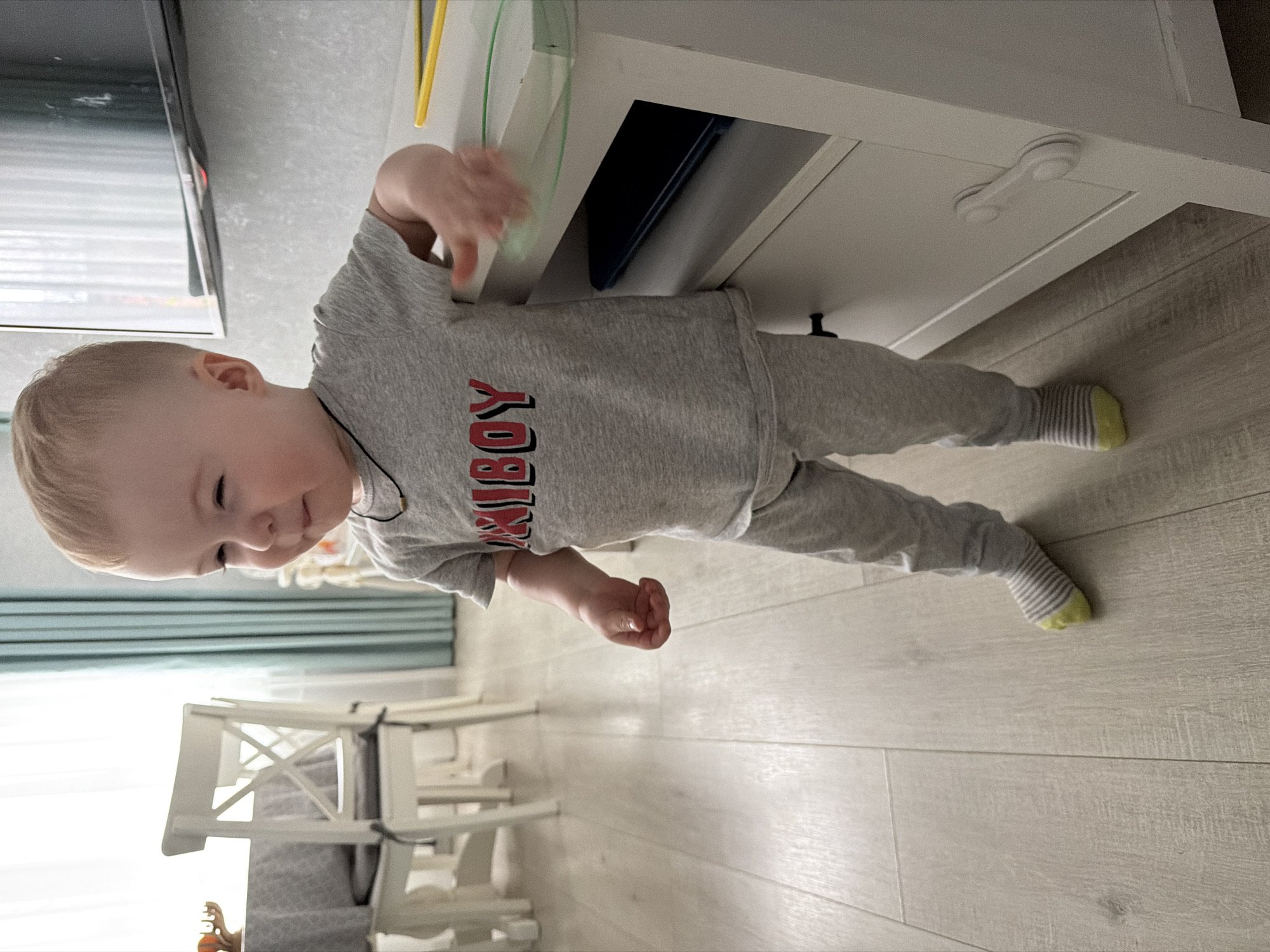 A young child is captured in an indoor, brightly lit setting, likely a home in Votkinsk, Russia, during the daytime. The child, with light blonde hair, is standing upright and appears to be a toddler, dressed in a grey long-sleeved shirt with the red word "BOY" prominently displayed, preceded by some lighter grey letters that are partially obscured (possibly "XIBOY" or "PIBOY"). They also wear matching grey pants and white and grey striped socks with a yellow heel/toe. A dark cord or necklace is visible around the child's neck.
The child is seen looking down and to their left with a gentle, curious expression, possibly a slight smile, while interacting with a white cabinet or dresser. Their right hand is reaching towards or resting on the top edge of the cabinet. The cabinet features a visible white child safety lock on one of its drawers. Inside an open compartment of the cabinet, a dark blue object, possibly a tablet or book, is visible, and above it, a yellow and green object.
The background shows light-colored walls, light wood laminate flooring, and light blue/green curtains partially covering a window. A television screen is partially visible on the left, and a white chair or high chair with a cross-back design is seen in the lower left background, suggesting a living or dining area. The overall scene conveys a sense of innocent exploration and domestic comfort.