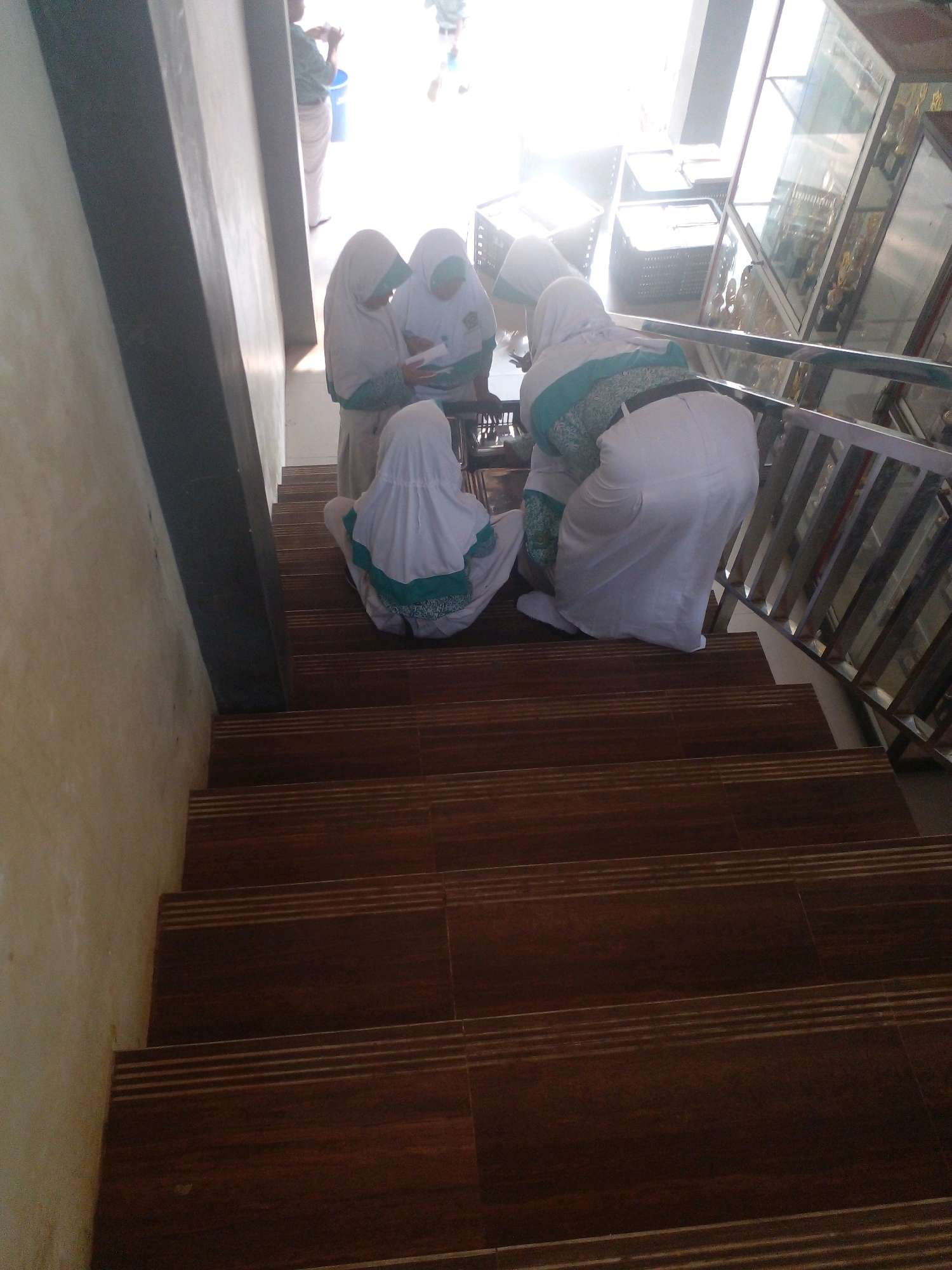 The image shows four young women, likely students, dressed in white hijabs and school uniforms, gathered on a staircase in a building. One woman is standing and holding what appears to be a notebook. Two other women are kneeling beside her, and a fourth woman is kneeling further down the stairs, facing away from the camera. The staircase has dark brown tiled steps with lighter brown horizontal lines. A metal railing runs along the right side of the stairs, with a display case filled with trophies visible behind it. The building's wall on the left is light beige with a dark grey beam. The scene suggests a casual moment of interaction between the students, possibly during a break or in transit. The location of Banyuputih, Indonesia, is noted, implying this is likely a school or public building within that region.