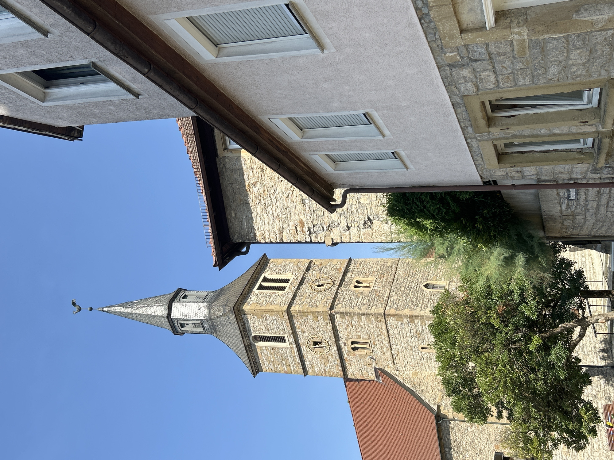 The image shows a view of the St. Nikolaus Church in Bad Friedrichshall, Germany. The church features a tall, stone tower with a steeply pitched, gray, slate-covered roof topped by a weather vane shaped like a rooster. The tower has several arched windows and what appear to be clock faces on at least two sides. Adjacent to the church is a building with a light-colored, textured facade and stone foundations, featuring several windows with white frames and roller shutters. Another stone building with a red tile roof is visible behind the church tower. Lush green trees and shrubs are present in the foreground and to the side of the buildings. The sky is clear and blue.