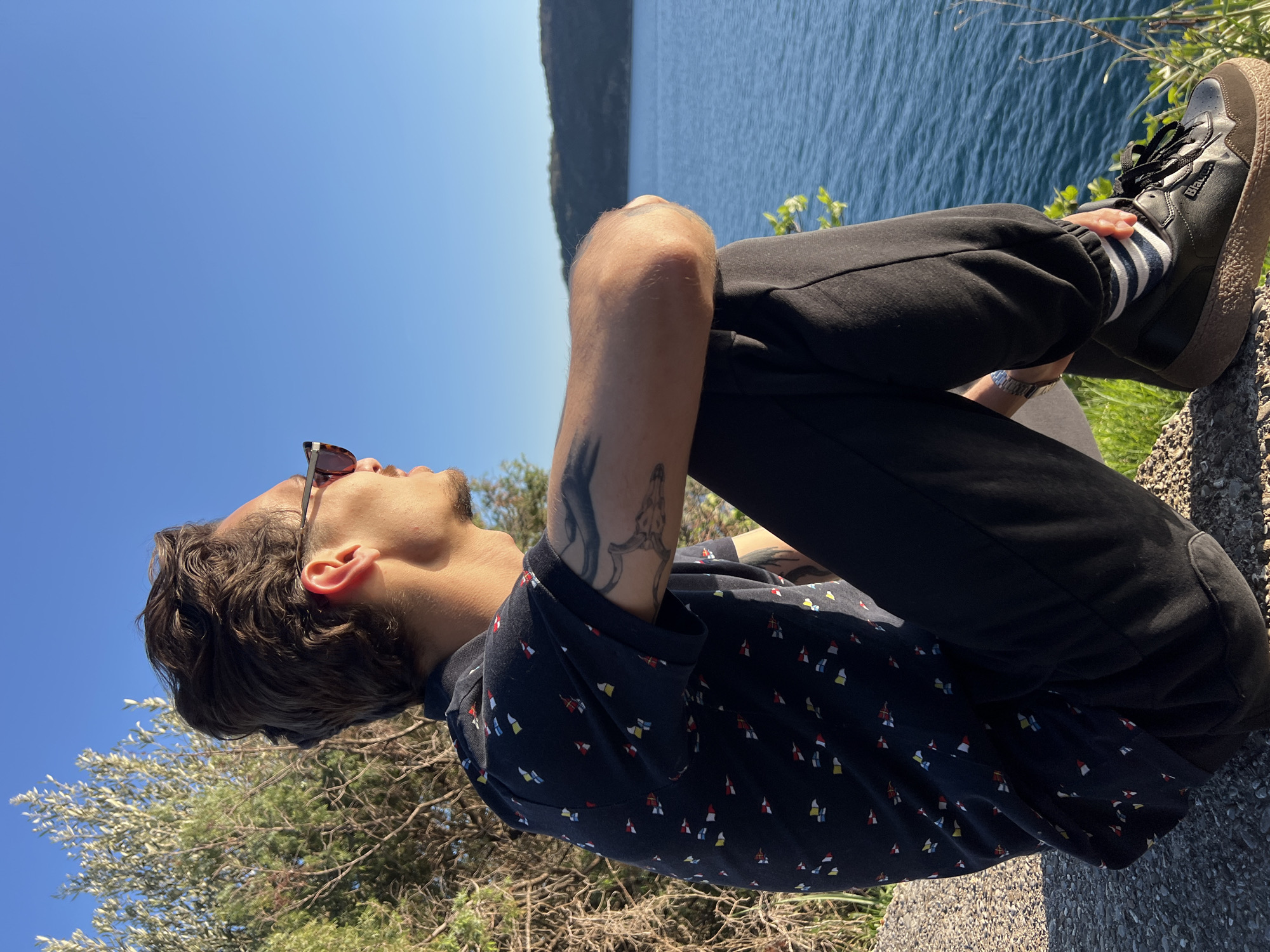 A person is sitting on a rock ledge in an outdoor setting, possibly in Budva, Montenegro, given the coastal and hilly landscape. The person is wearing sunglasses, a dark-colored shirt with a colorful pattern, black pants, black socks with white stripes, and black sneakers with brown soles. Their arms are wrapped around their legs, and they are looking up towards the sky. A tattoo is visible on their left arm. In the background, there's a body of water, likely the Adriatic Sea, with a land mass visible across the water. The sky is clear and blue. Some green foliage and vegetation are present near the ledge where the person is sitting. The image appears to be taken during the daytime with bright sunlight.