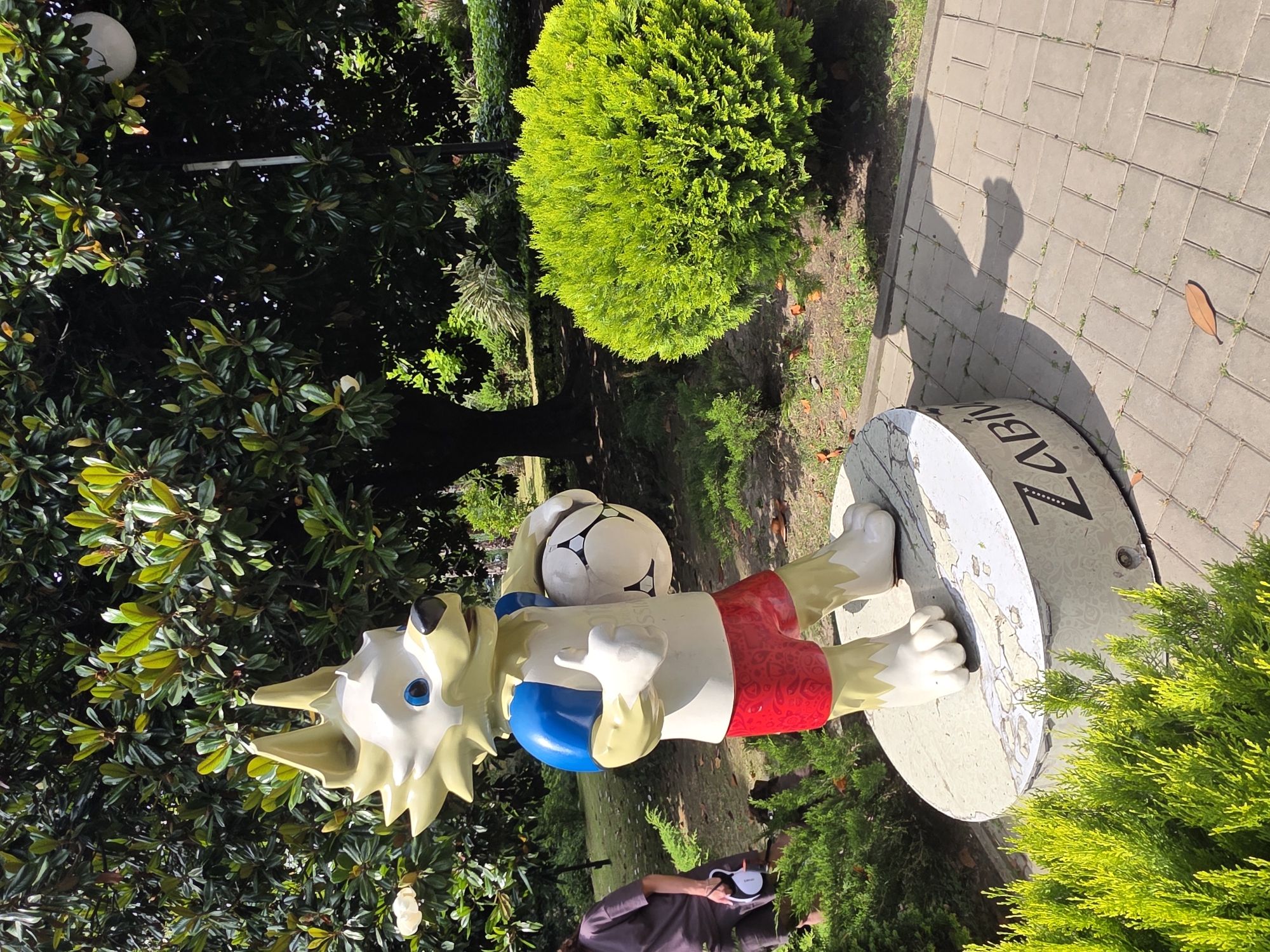 This is a vertically oriented photograph of a statue of Zabivaka, the wolf mascot for the 2018 FIFA World Cup, standing on a circular base. The statue is depicted with yellow fur, blue eyes, red shorts with a white pattern, and a blue and white soccer ball. The statue is positioned on a paved area with a shadow cast on it.
In the background, there are lush green trees and bushes, including a large, rounded green shrub. The setting appears to be an outdoor park or garden. The sunlight suggests it is daytime. The overall atmosphere is bright and cheerful, consistent with a public display of a popular sports mascot.
The base of the statue has visible text that reads "ZABI" followed by a series of dots and then "KA". The paving stones are rectangular and laid in a herringbone pattern. A person wearing a dark top is partially visible in the lower right portion of the frame, appearing to be holding a dog leash. The location context is Sochi, Russia.