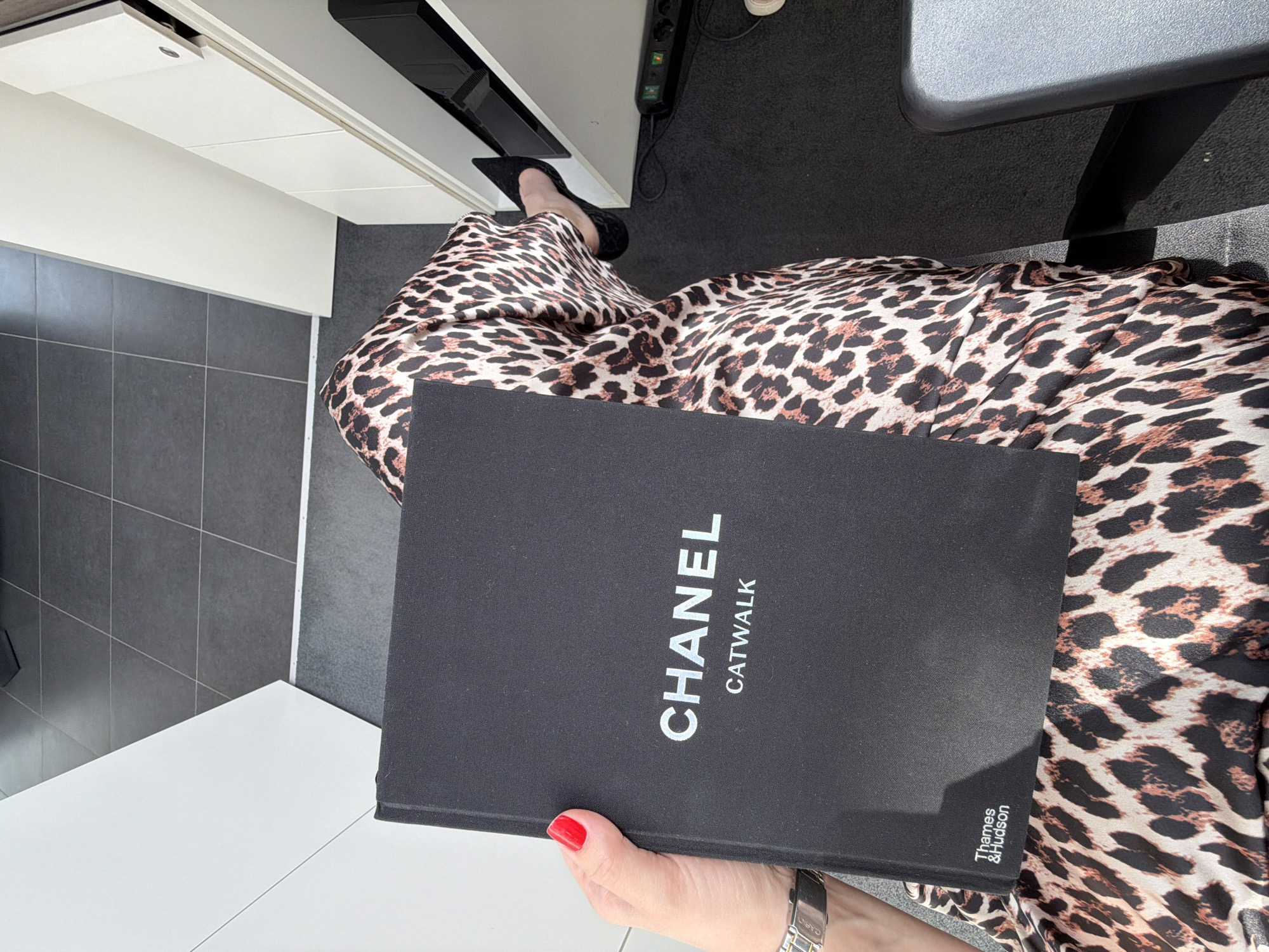 A high-angle shot captures a person seated at a desk or table, clad in leopard-print pants and black high heels. The person holds a black book titled "CHANEL CATWALK" with "Thames & Hudson" printed near the bottom right corner. The person has red nail polish on their nails and wears a watch on their left wrist. The setting appears to be an office space or home office, with a white desk or countertop visible to the left, gray tile flooring, and a black computer monitor or device above. There's also a black power strip with outlets visible next to the desk. The environment is well-lit. There is nothing to indicate this image was taken in the city of Izhevsk, Russia.