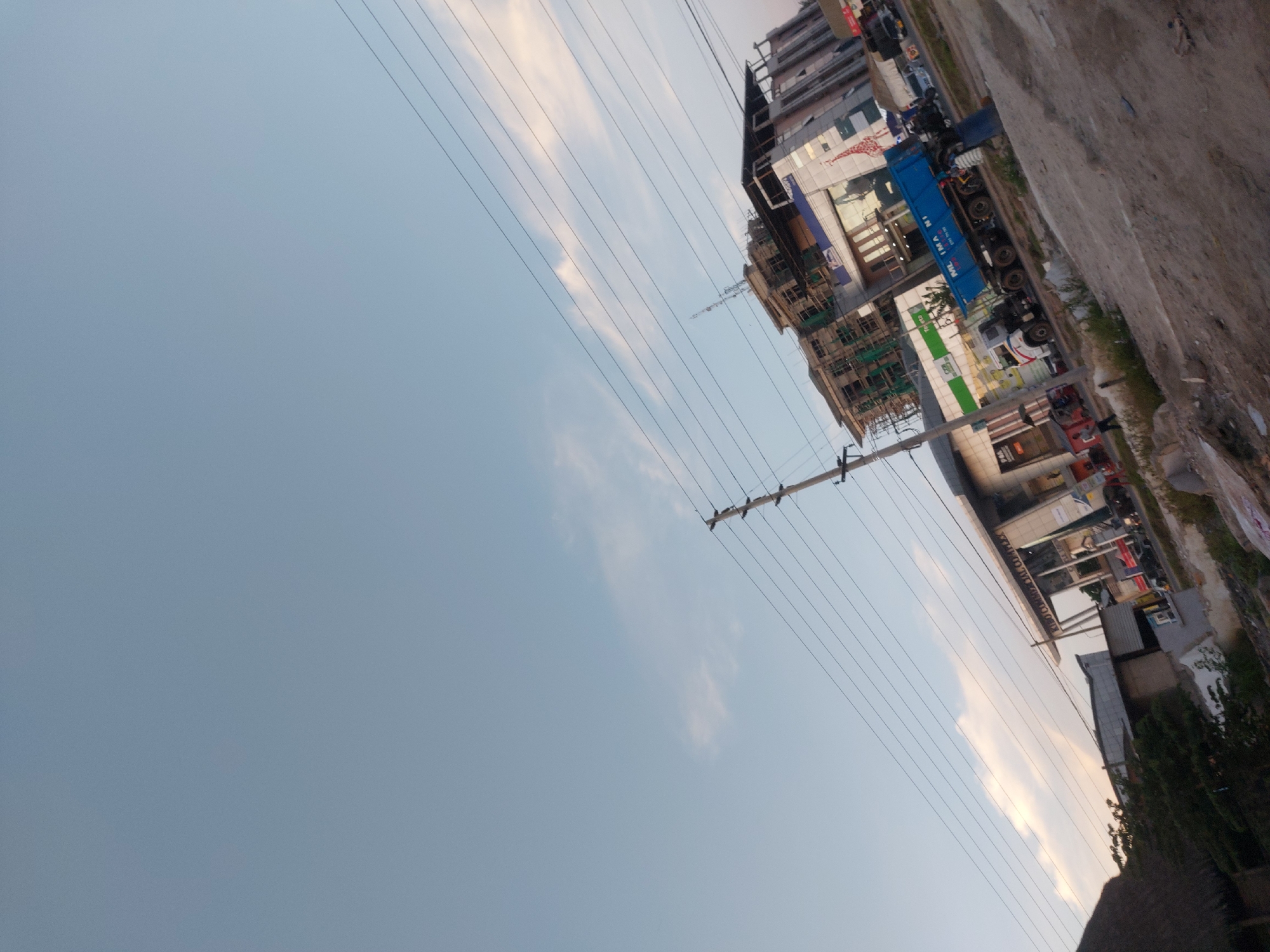 The image depicts a street scene in Dar es-Salaam, Tanzania, looking up at an angle. The sky occupies a large portion of the frame on the left. A utility pole with power lines runs diagonally across the image. On the right side are buildings, some under construction, and a blue truck. There are parked cars and motorcycles along the street. The ground consists of a concrete walkway with some greenery.