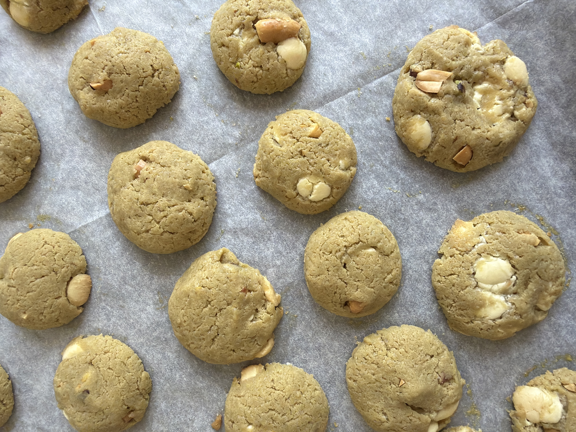 The image displays a close-up, overhead view of several freshly baked cookies arranged on a sheet of parchment paper. The cookies are round and have a light brown, slightly textured surface, suggesting they are likely made with ingredients such as butter, flour, and sugar. Some of the cookies appear to have chunks of white chocolate or nuts embedded within them, adding visual interest and suggesting a variety of ingredients. The parchment paper is light gray and slightly creased, indicating it has been used for baking. The lighting is bright and even, casting subtle shadows that highlight the shape and texture of the cookies. There is no indication of a specific location in Tel-Aviv, Israel, as the scene is confined to the baking sheet and cookies. The overall impression is one of homemade baked goods, ready to be enjoyed.