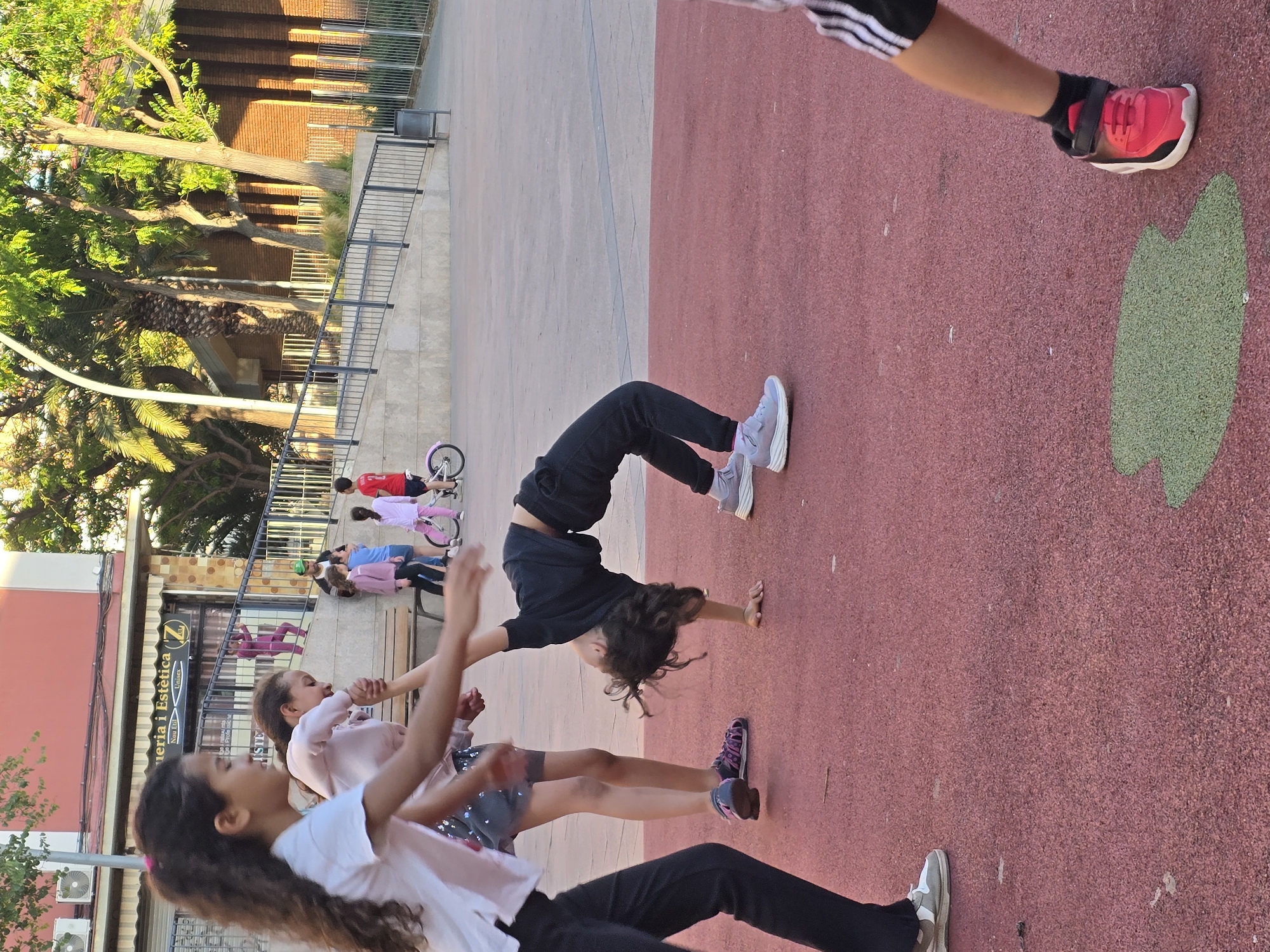 The image depicts a group of children on a playground in Cornellà De Llobregat, Spain. At least three children are visible in the foreground, one of whom is doing a backbend. Other children are visible in the background, some on bicycles. The playground has a red surface. Behind the playground is a building with shops. Trees are also visible in the background.