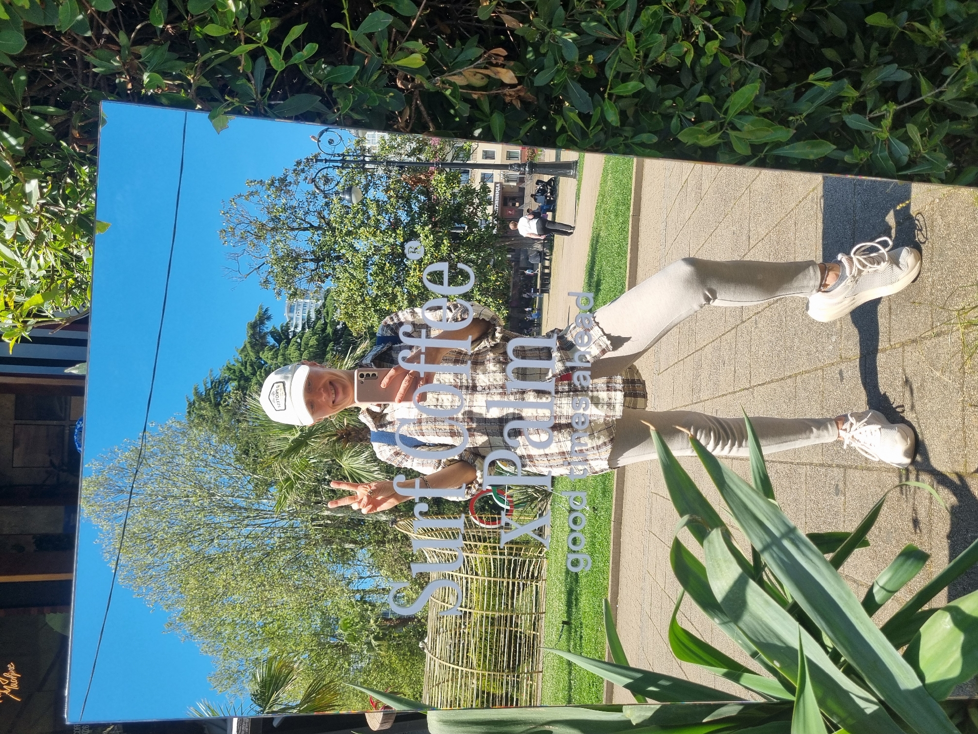 A full shot in vertical orientation captures a person's reflection in a mirror outdoors. The person is wearing a patterned shirt, light-colored pants, white sneakers, and a white baseball cap. They are holding a smartphone and making a peace sign.
The mirror reflects a clear blue sky, trees, and a building in the background. The mirror is positioned among plants, including shrubs at the top and large, spiky-leaved plants at the bottom. There's also what appears to be a decorative bamboo structure in the reflection.
The scene is set on a paved area, possibly a sidewalk or plaza. In the background of the mirror's reflection, other people can be seen walking or sitting on benches near old-fashioned lampposts, which indicate the location to be in a park. A building can be glimpsed in the top of the mirror.
In summary, the image shows a person taking a selfie in a mirror placed in an outdoor park or plaza setting.