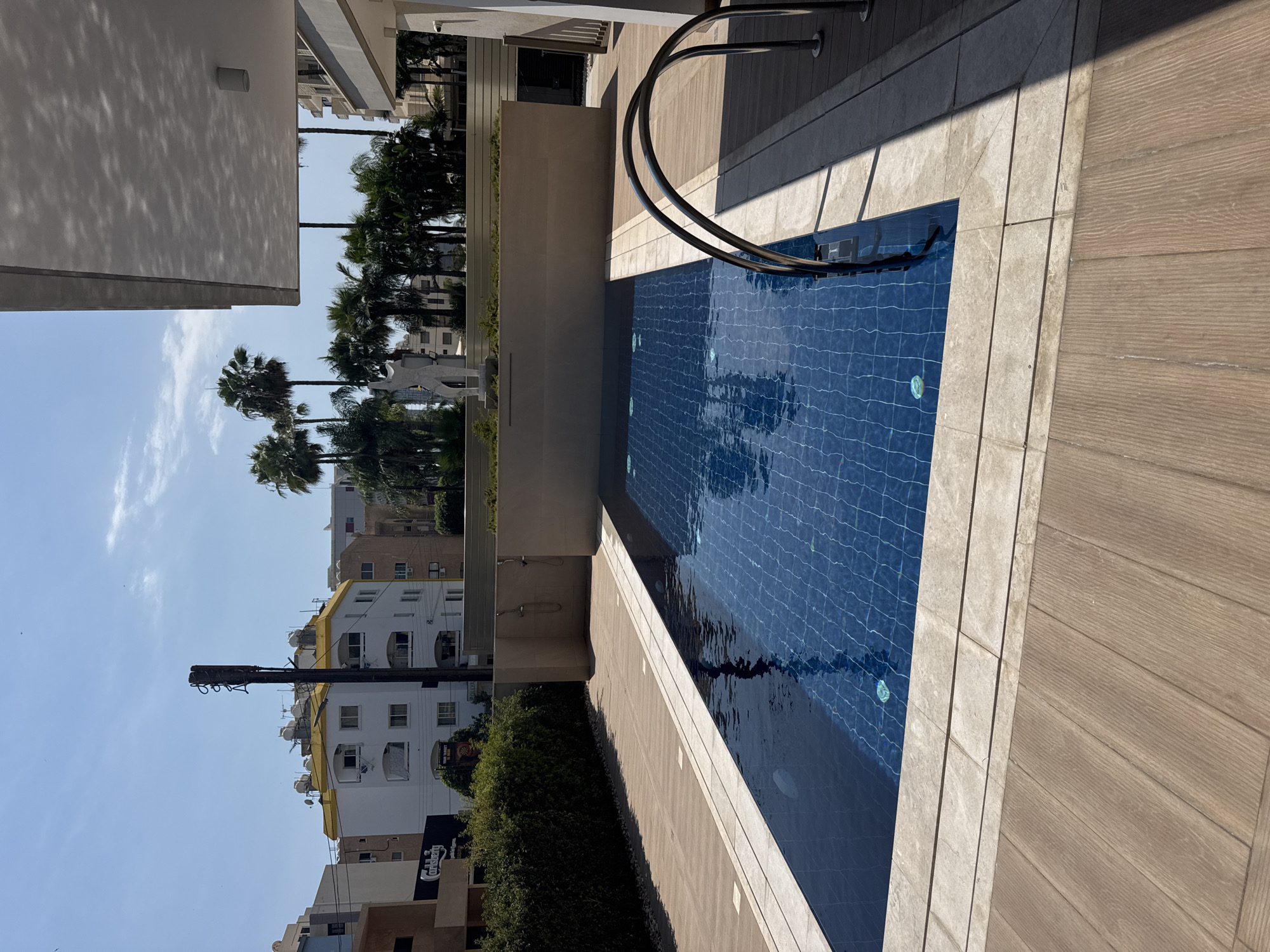 An outdoor swimming pool with a tiled blue bottom is situated on a paved area of light-colored tiles and wooden planks. The pool has chrome handrails and is surrounded by a beige stone border. Beyond the pool area, a low beige wall is topped with greenery and a white abstract sculpture. In the background, a cluster of palm trees stands against a bright blue sky. Further back, a street scene is visible, featuring a white building with a yellow roof, other apartment buildings, and a utility pole. The overall environment suggests a sunny, urban setting in Germasogeia Municipality, Cyprus. There are no people visible in the image.