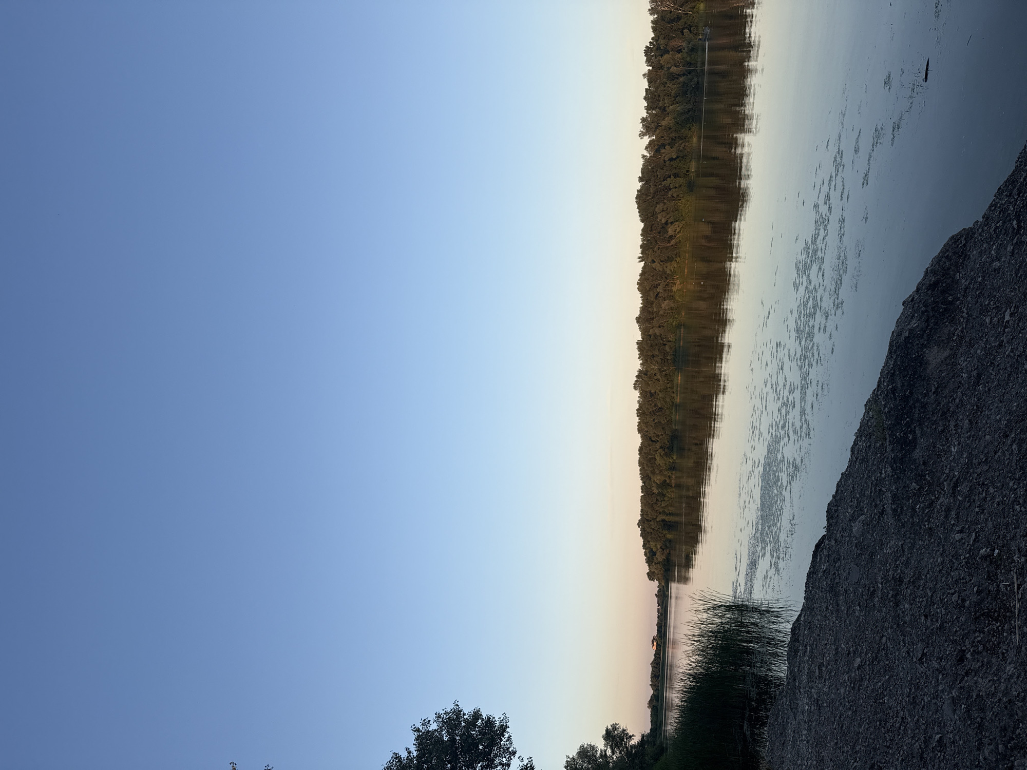 This media file depicts a tranquil natural scene by a body of water, likely a river given the context of Dnipro, Ukraine. The image is oriented vertically, suggesting it was captured with a phone.
The primary subjects are the serene water, a dense line of trees forming the opposite bank, and a rocky, gravelly embankment in the foreground. The water's surface is calm, reflecting the twilight sky and the treeline. Scattered patches of aquatic vegetation dot the water. Tall reeds or marsh grass grow along the near shore, transitioning into the rocky terrain. The trees on the far bank are lush and green, forming a continuous line that stretches horizontally across the frame.
The time of day appears to be either early morning or late evening, during twilight. The sky transitions from a clear, pale blue at the top to softer hues of pink and yellow near the horizon, indicating the sun is low in the sky. The weather seems calm and clear, with no visible clouds or wind.
There is no discernible activity or event taking place; the scene is one of natural stillness and peace. A very faint silhouette of what might be a building or structure can be seen on the far bank, near the water's edge, among the trees.
The overall mood is one of serenity and quietude. The combination of the calm water, the fading light, and the natural landscape creates a peaceful atmosphere. There is no visible text in the image.