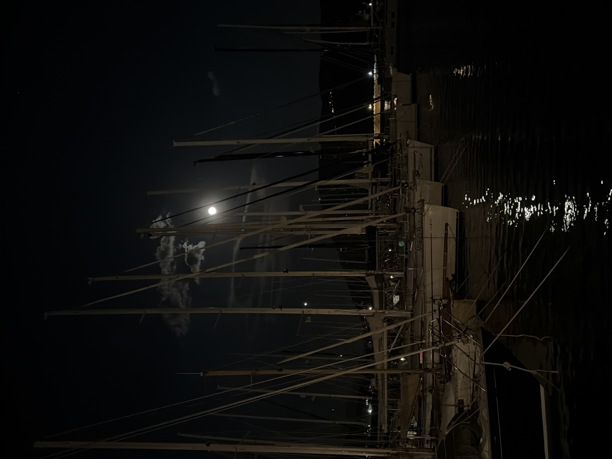 The image captures a nighttime scene in Meljine, Montenegro, featuring a row of docked sailboats with their masts reaching towards the sky. The moon is visible through the masts, partially obscured by thin clouds. Reflections of light can be seen shimmering on the water's surface. Dockside structures and lights are dimly visible in the background.