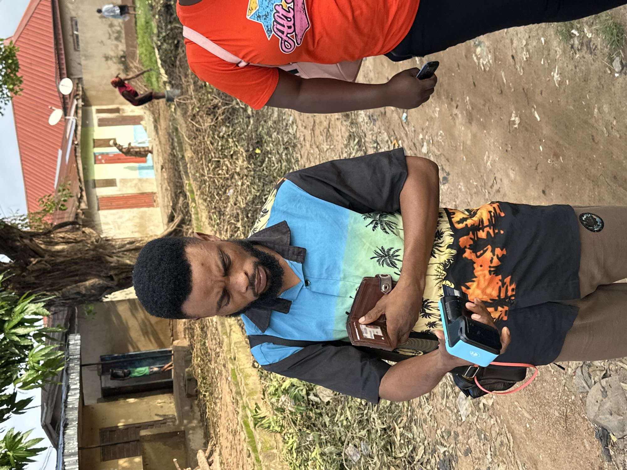 A medium shot captures a Black man in Narayi, Nigeria, standing outdoors and looking down at a brown leather wallet he holds open in his left hand. He wears a collared, short-sleeved shirt with a blue top half and a black and orange palm tree print on the bottom half, along with brown pants. His right hand is holding a small, blue and black electronic device, possibly a card reader. A black bag with a red strap is slung over his left shoulder. In the background, a woman in an orange t-shirt is partially visible on the right side of the frame. The environment appears to be a dirt road lined with dry vegetation and debris. A few buildings with corrugated metal and red roofs are visible further in the background, along with a large tree. The lighting suggests it is daytime.