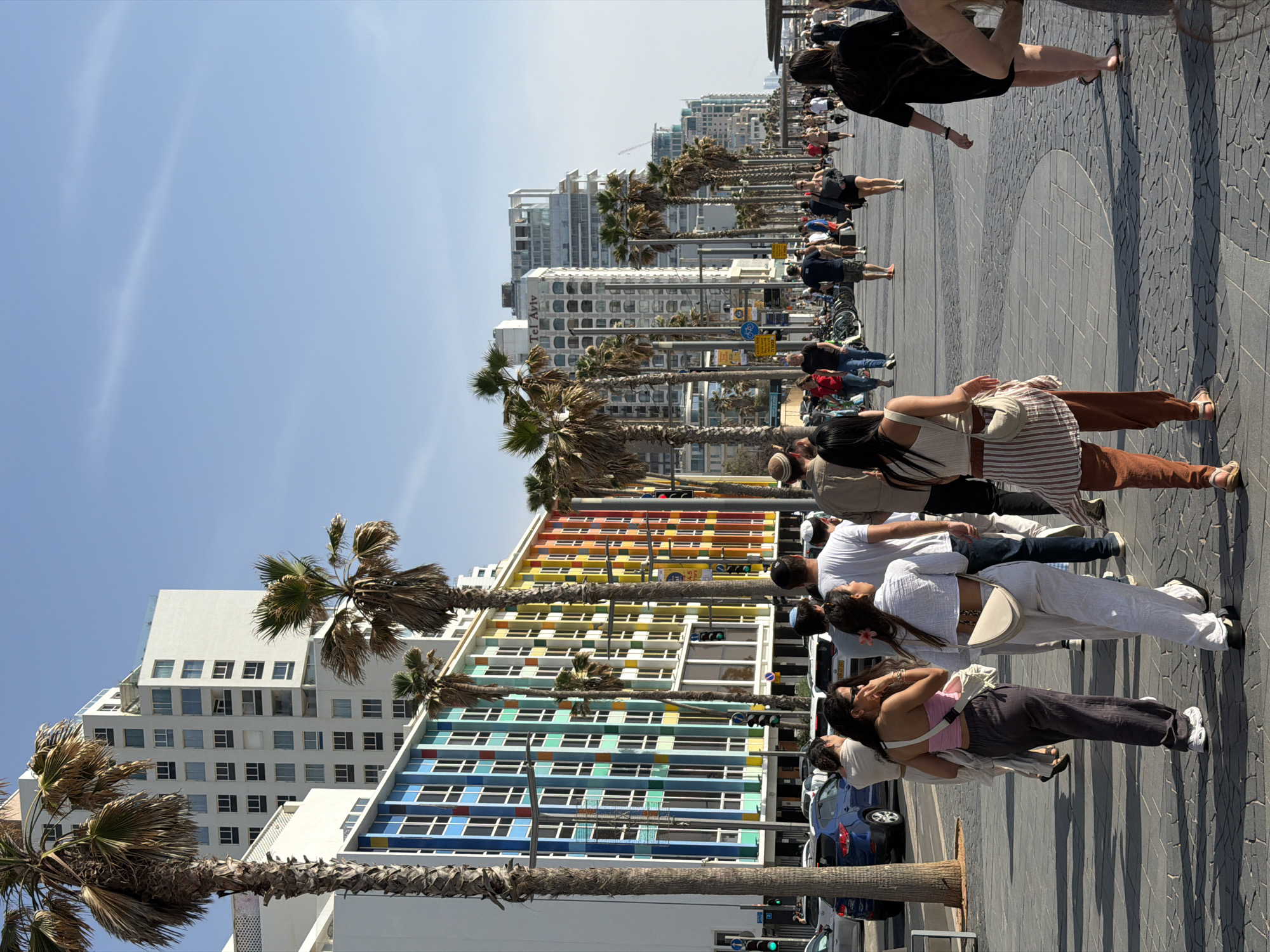 The photograph depicts a street scene in Tel Aviv, Israel, likely taken along the beachfront promenade. A line of people, likely tourists or locals, are observing something of interest, their attention directed towards an unseen subject to the right of the frame. The streetscape includes palm trees lining the walkway and various buildings, including a distinctive multicolored building with numerous windows, adding a playful element to the cityscape. The individuals in the scene are dressed in casual attire, reflecting a relaxed, daytime atmosphere. Cars are visible on the street parallel to the walkway, indicating an urban setting. The environment is bright and sunny, suggesting a pleasant day by the Mediterranean Sea.