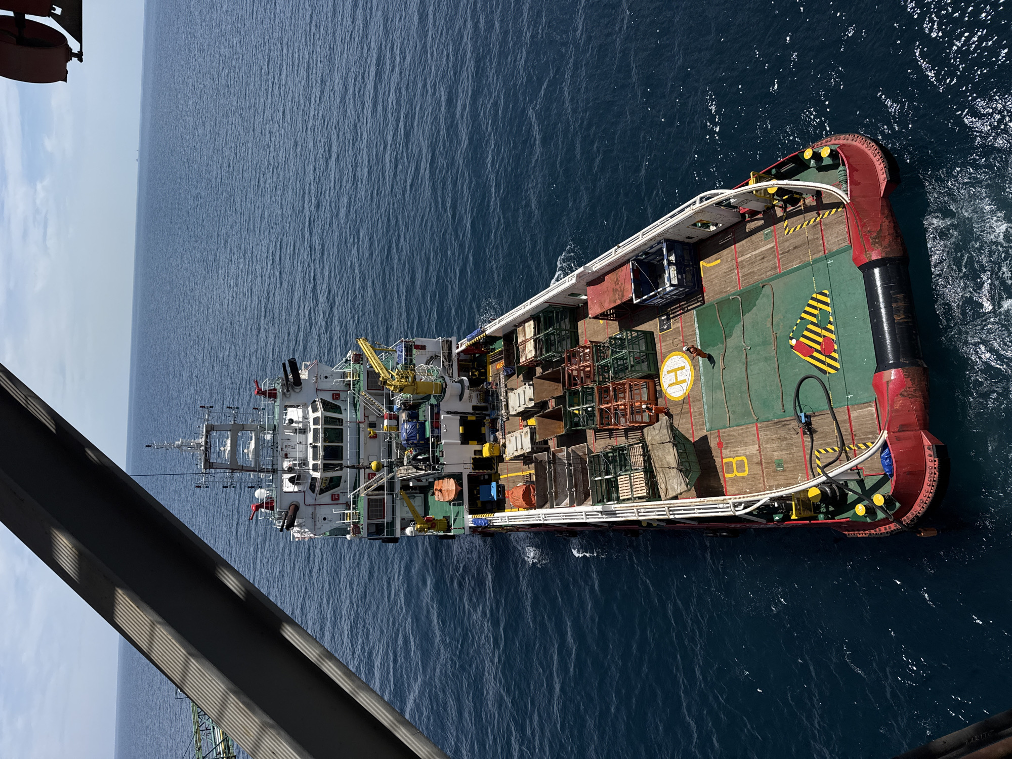 An aerial, high-angle, full shot captures a red and white supply ship sailing on a calm, blue sea under a partly cloudy sky. The ship is laden with various cargo, including wooden crates, metal cages, and what appear to be large pipes or tanks. A helideck is visible on the aft deck. The foreground is partially obscured by a large, grey, angular structure, possibly part of a larger vessel or crane, casting diagonal shadows across the scene. There is one person visible on the deck wearing an orange uniform. The overall environment is maritime and industrial. The location is unknown; it is not possible to determine the city of Unknown, Italy from the provided image.